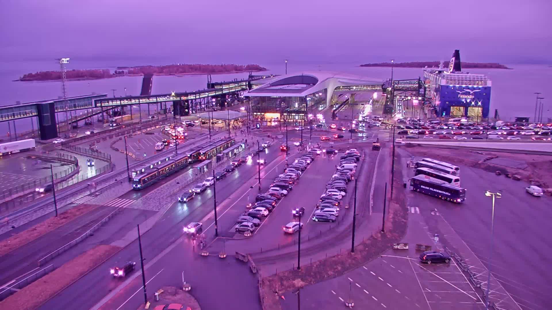 A ferry terminal at dusk, with a large ferry docked, numerous cars parked and driving around, a tram, buses, and a pedestrian walkway leading to the terminal.
