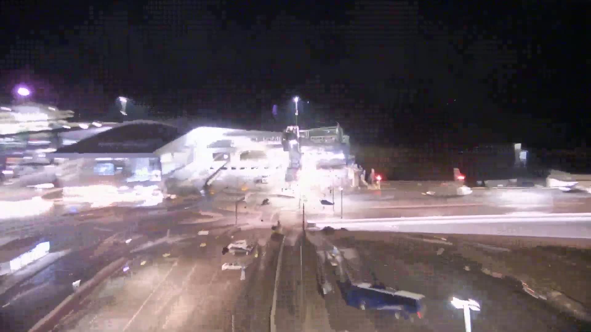 At night, a large building is illuminated, with vehicles and machinery surrounding it in a wet, dark area.