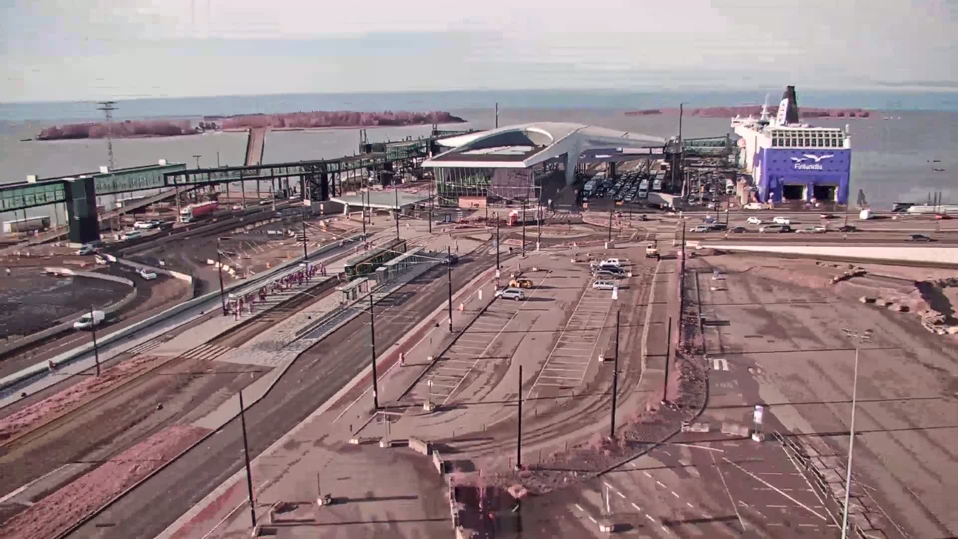 An aerial view shows a ferry terminal under construction on a partly cloudy day, with a ferry docked and vehicles parked nearby.