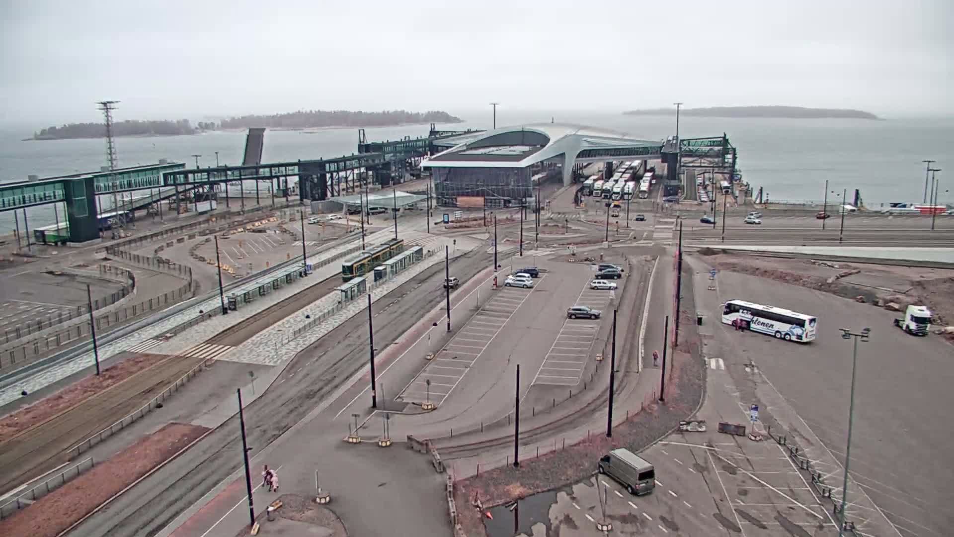 An aerial view of a ferry terminal on an overcast day shows a tram, buses, cars, and a partially constructed area.