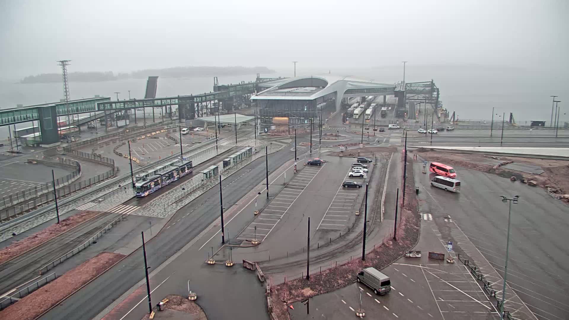 A ferry terminal on a foggy day with a tram, cars, and buses in the parking lot and on the road.
