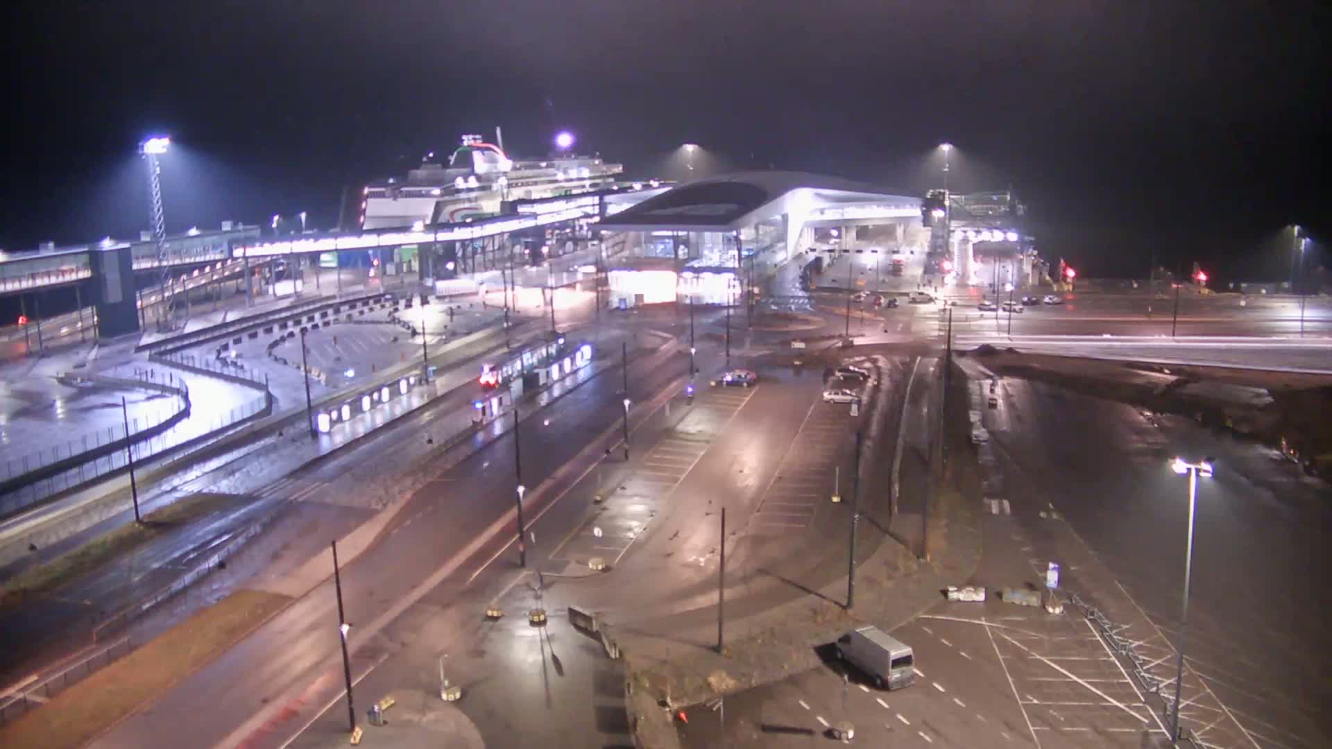 A large ferry is docked at a terminal at night in the rain, with vehicles and a tram visible in the surrounding area.
