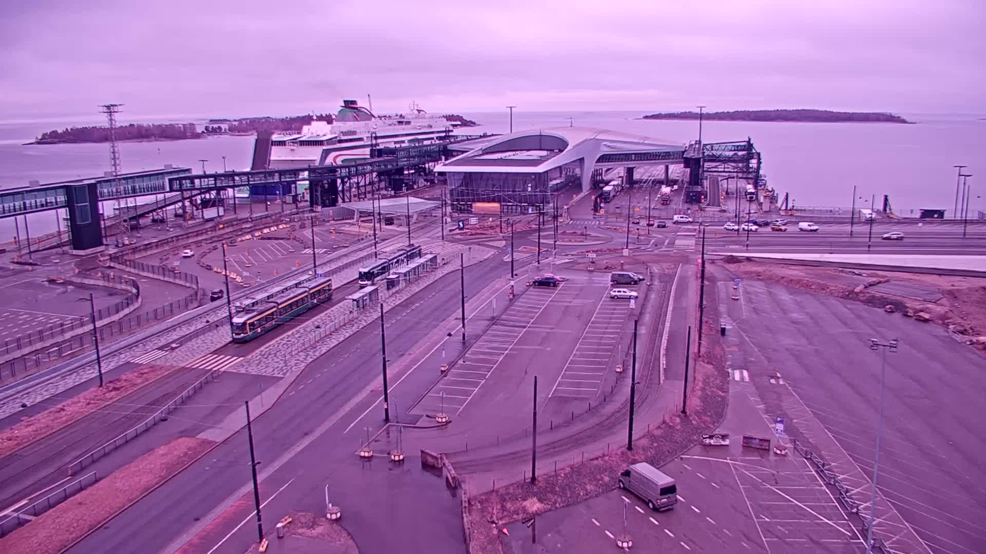 A ferry is docked at a terminal with a tram, cars, and a van in a parking lot under an overcast sky.