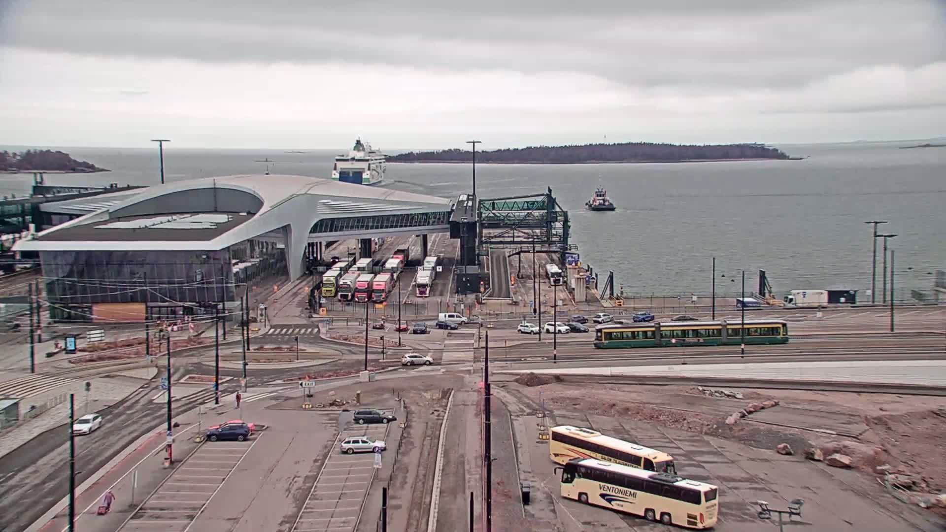 A ferry terminal with a large modern building, numerous buses and vehicles, a tram, a tugboat in the water, and a ferry in the distance under an overcast sky.