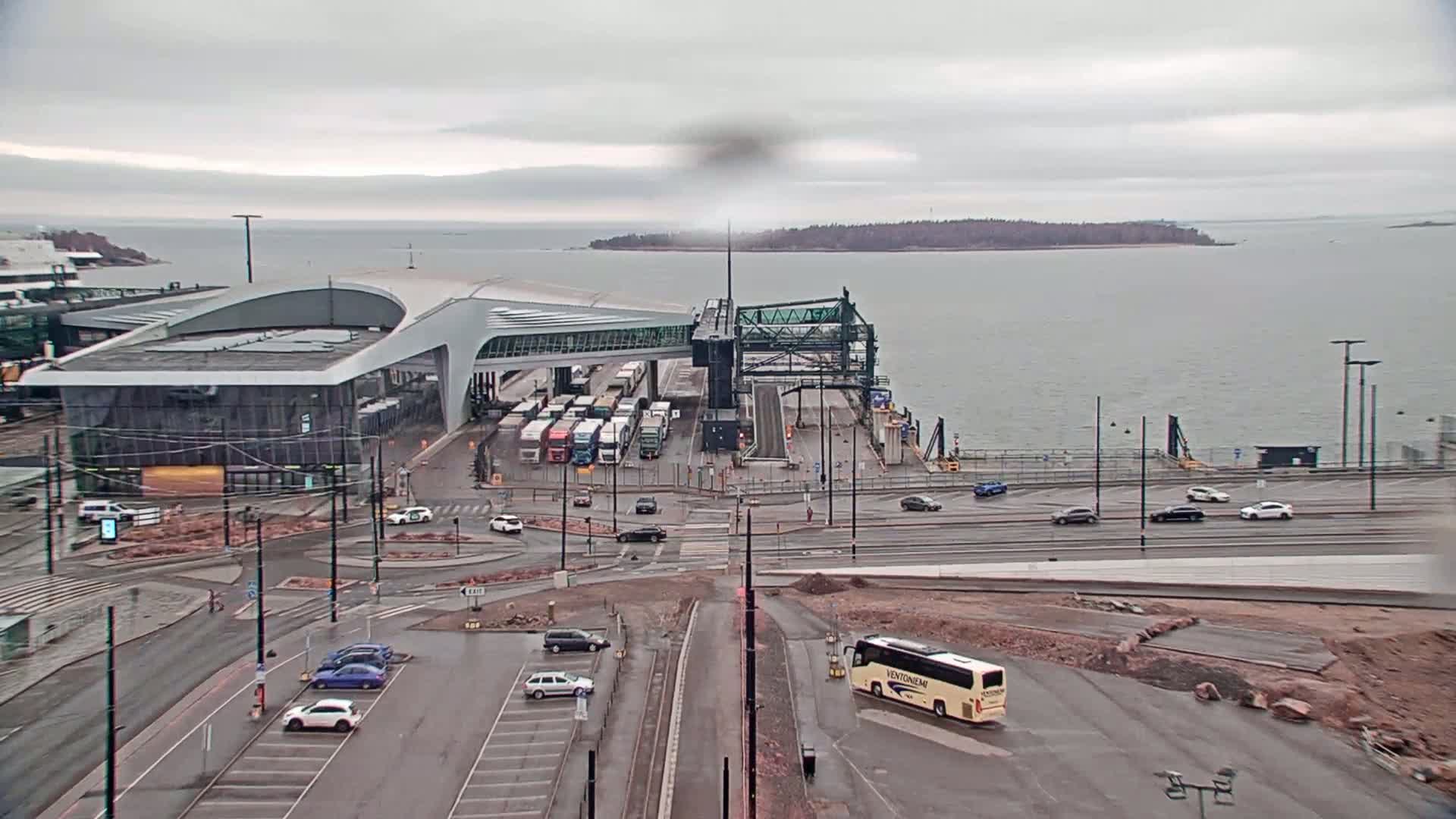 A ferry terminal with several vehicles parked and driving around it on an overcast day, next to a body of water.