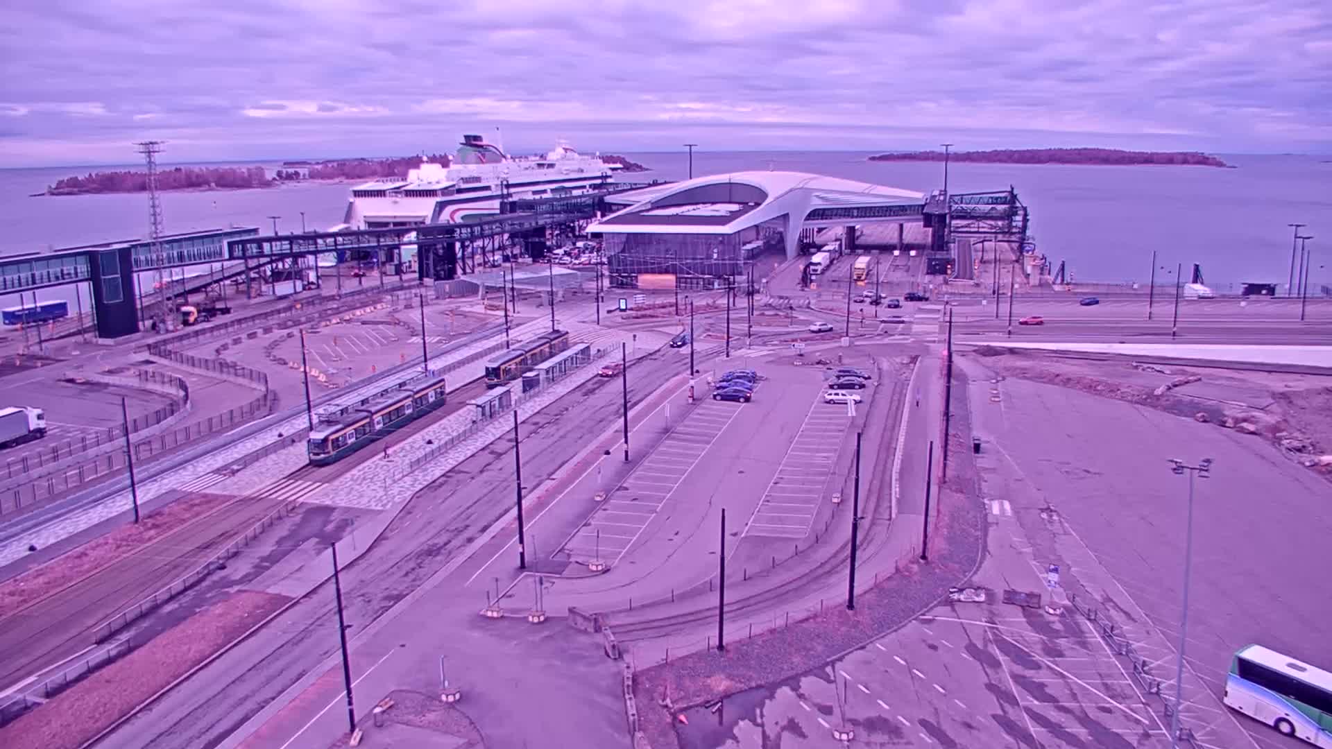 An aerial view shows a ferry terminal with a docked ferry, trams, cars in parking lots, and a calm body of water under an overcast sky.