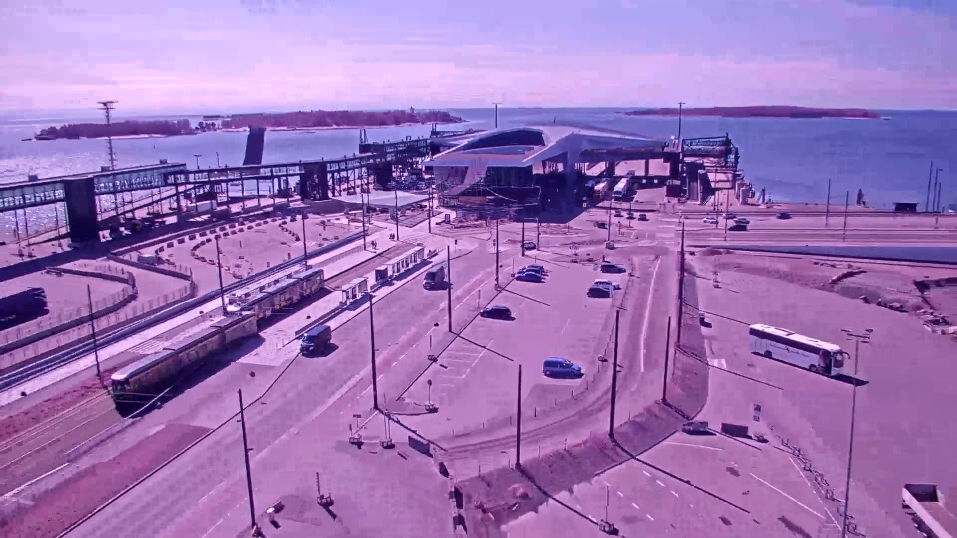 An aerial view shows a ferry terminal with a train on tracks, several vehicles in a parking lot, and a bus, all under a sunny sky.