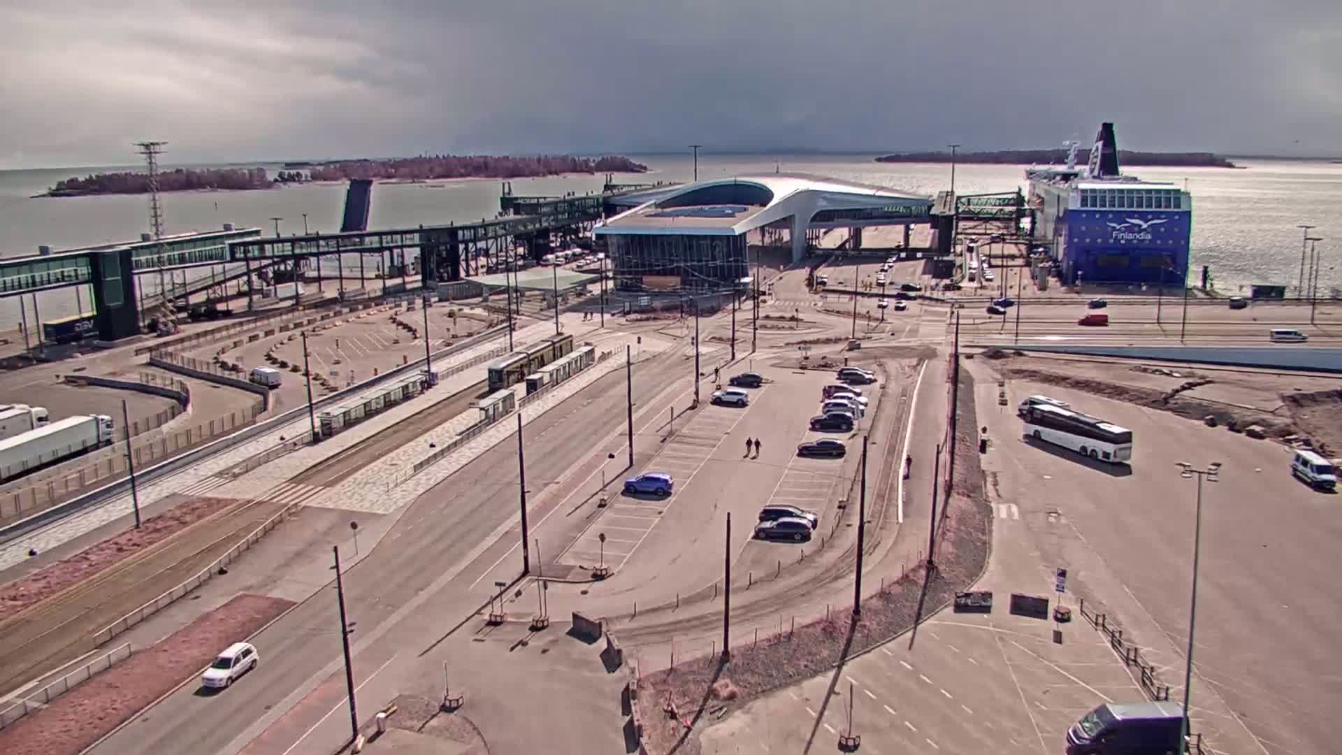 An aerial view of a ferry terminal on a partly cloudy day shows a large ferry docked, a modern terminal building, passenger walkways, parking lots with several vehicles, and a tram line.