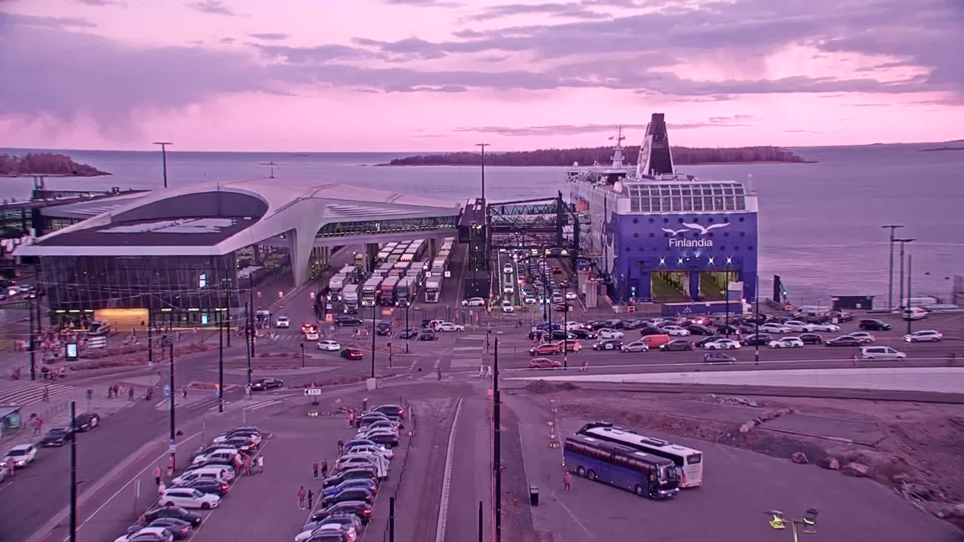 A large ferry is docked at a terminal, surrounded by numerous cars and trucks under a pinkish-purple sunset sky.