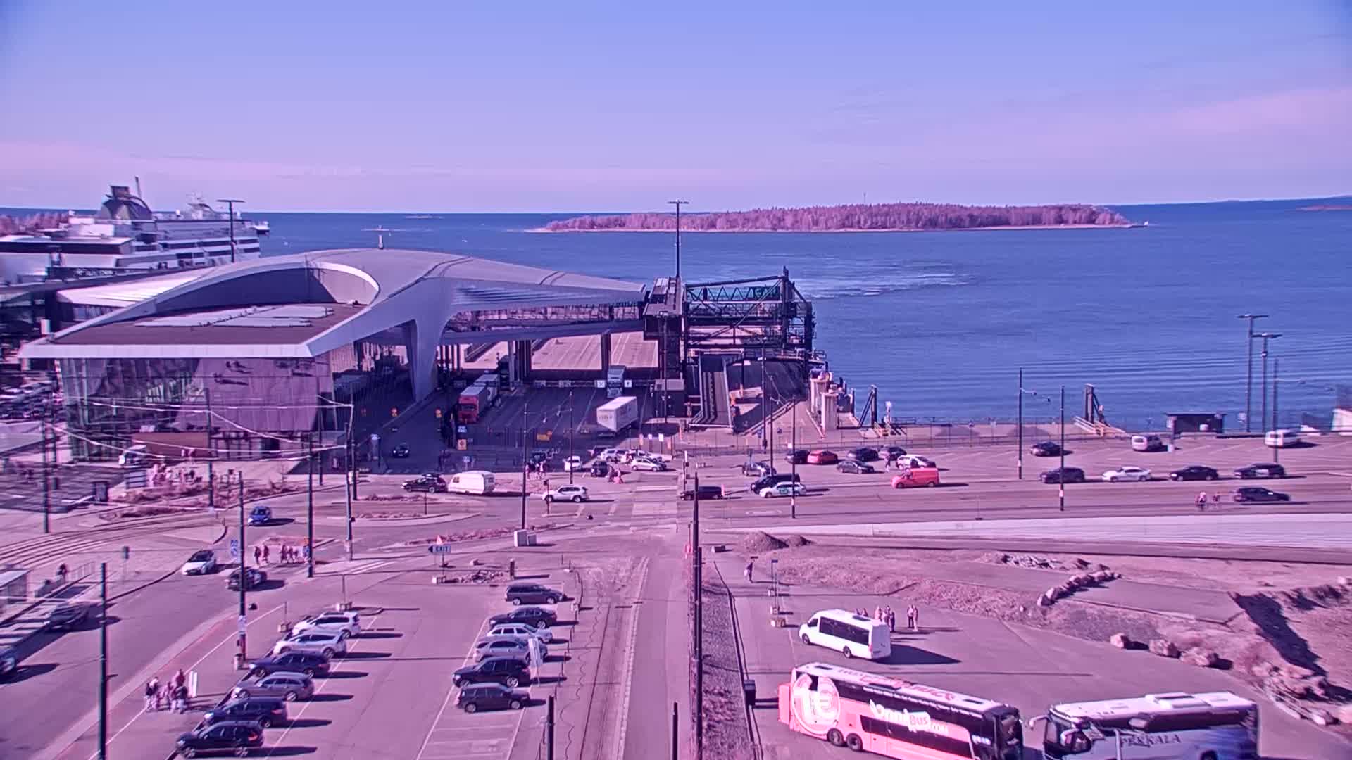 A ferry terminal with a large building and numerous vehicles, next to a body of water under a clear blue sky.
