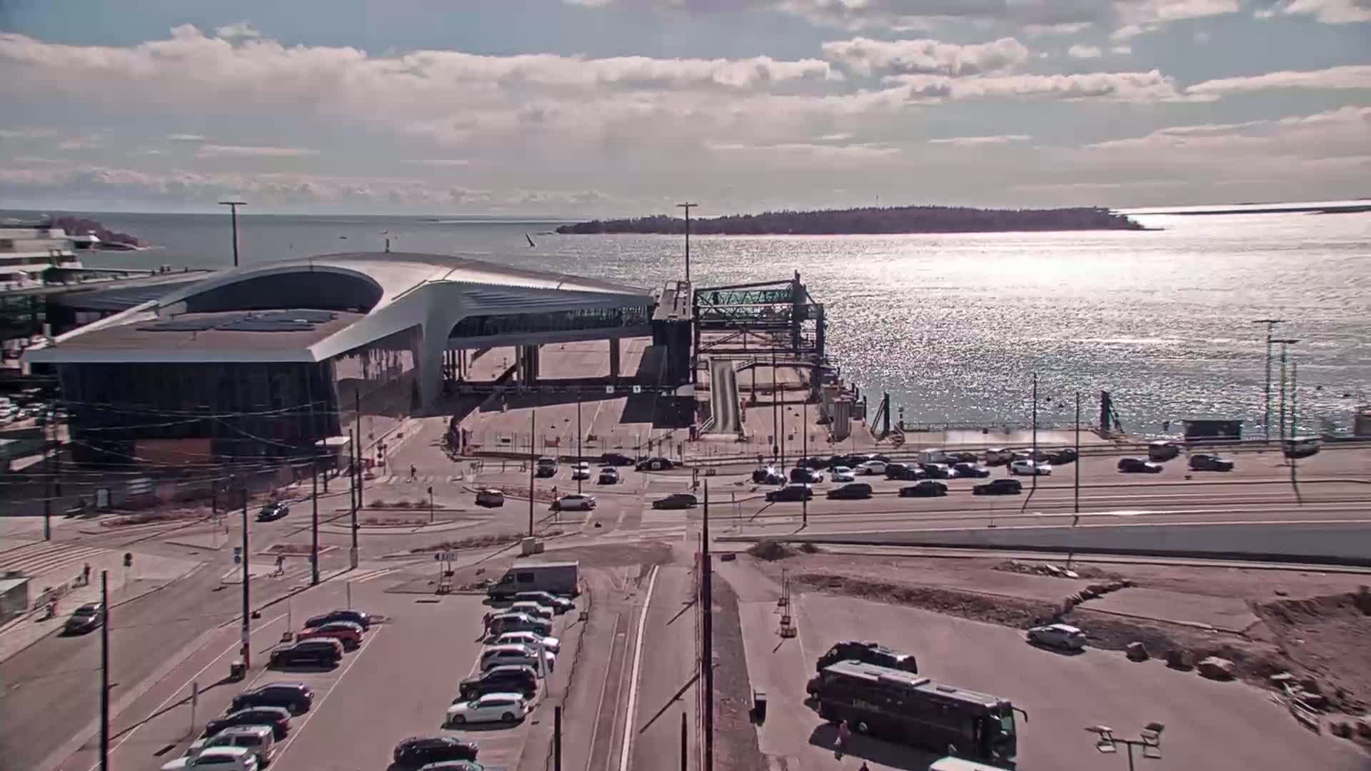 A ferry terminal with cars parked nearby overlooks a calm, sunlit body of water and a distant island under a partly cloudy sky.