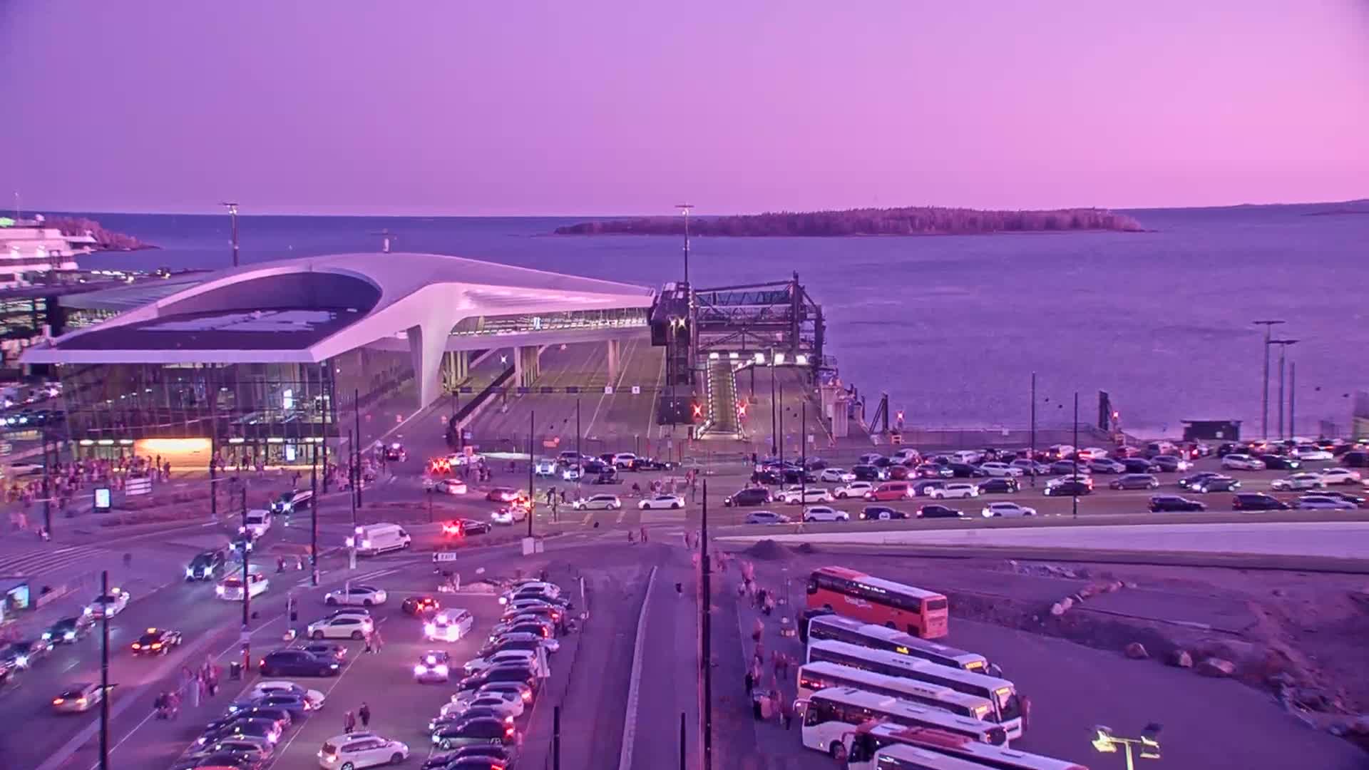 A ferry terminal with many cars and buses parked nearby under a purple twilight sky.