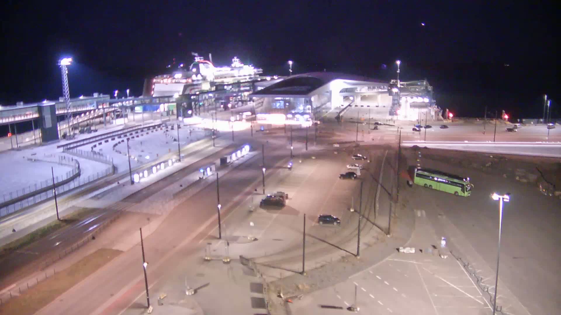 A large ferry is docked at a terminal at night, with cars and a bus parked in the surrounding lots, under clear skies.