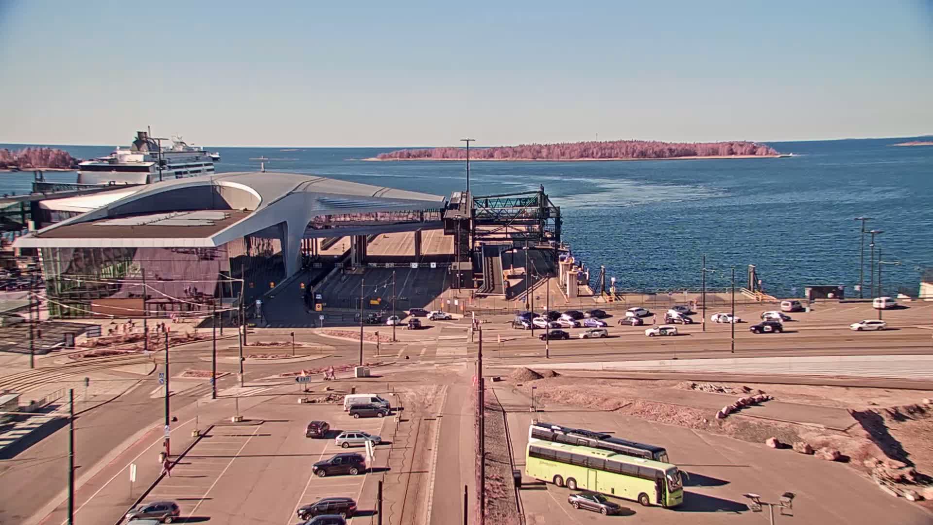 A ferry terminal with a modern building and several cars and buses is situated by a body of water on a sunny day.