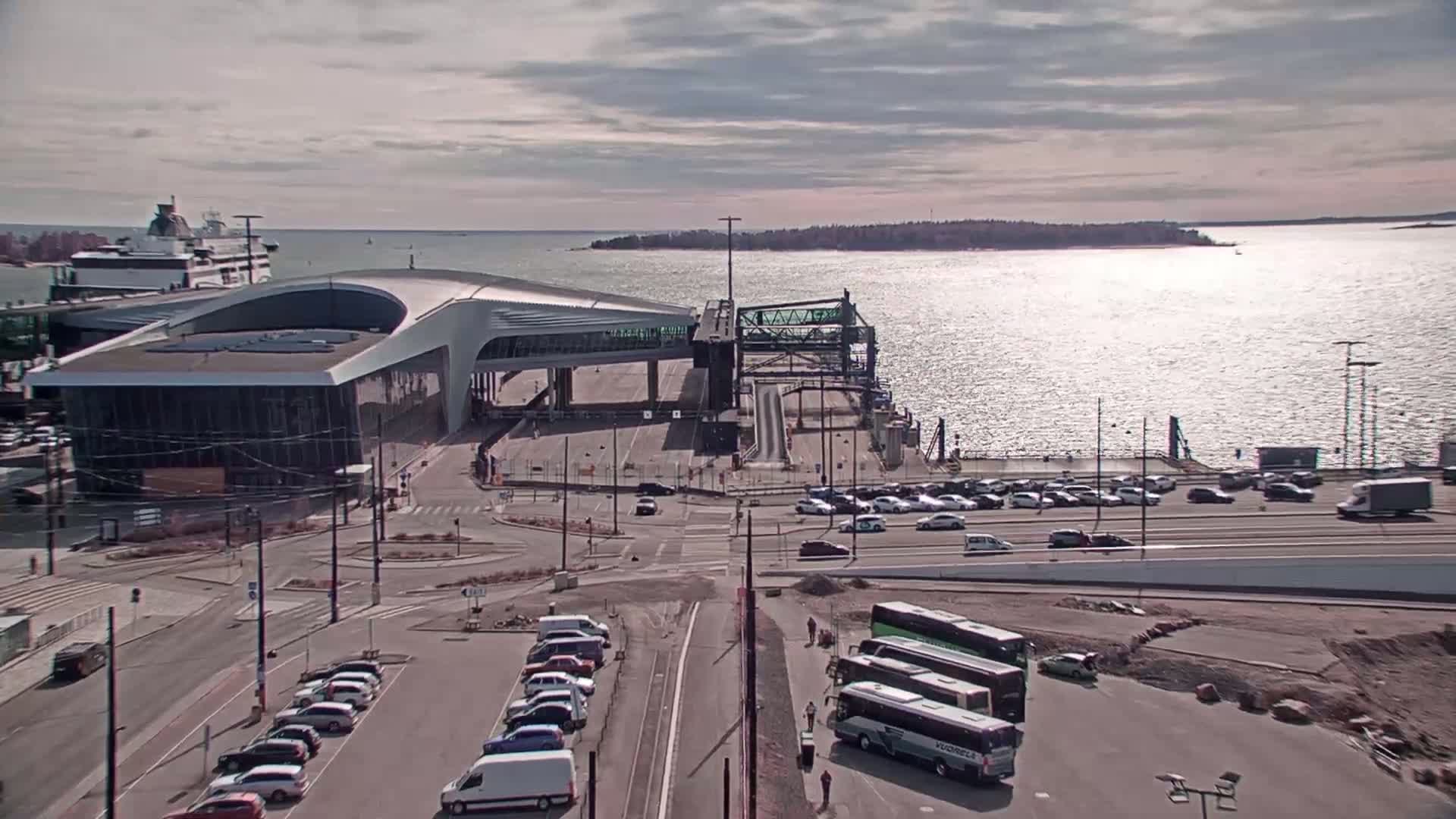 A ferry terminal with a large modern building, numerous cars and buses in parking areas, and a body of water with a distant island under a partly cloudy sky.