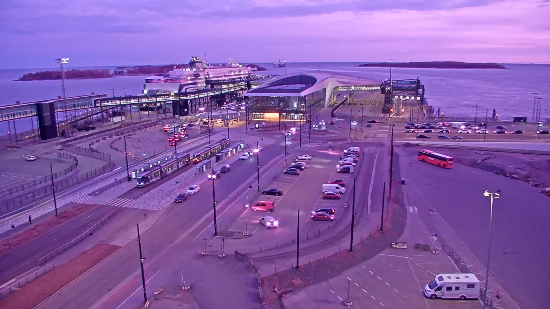 A ferry terminal at dusk with a ferry docked, a tram, numerous cars in a parking lot, and a bus, all under a purple twilight sky.
