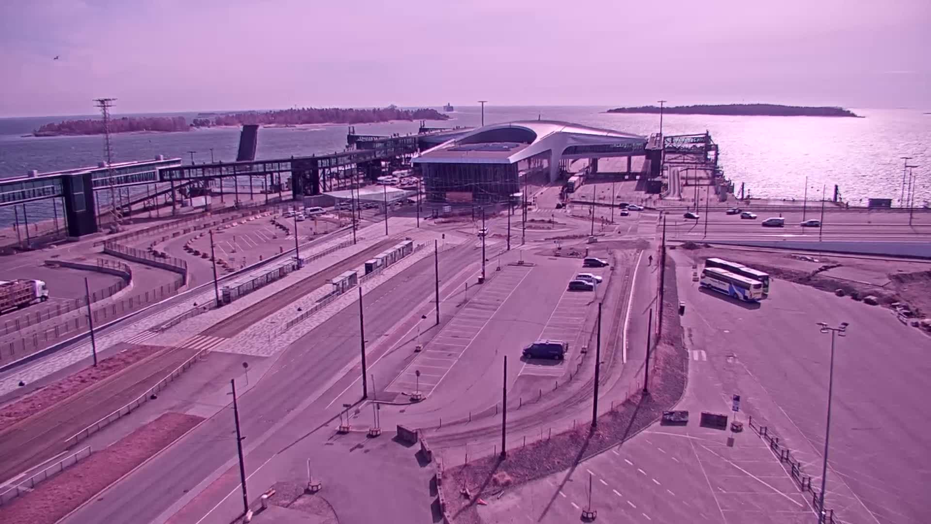An aerial view shows a ferry terminal with a modern passenger building, roadways, parking lots, and several vehicles including buses, on a sunny day by the water.
