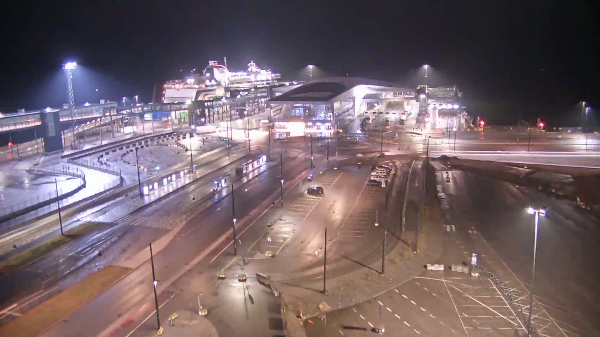 A large ferry is docked at a terminal at night in the rain, with empty parking lots and roadways surrounding it.