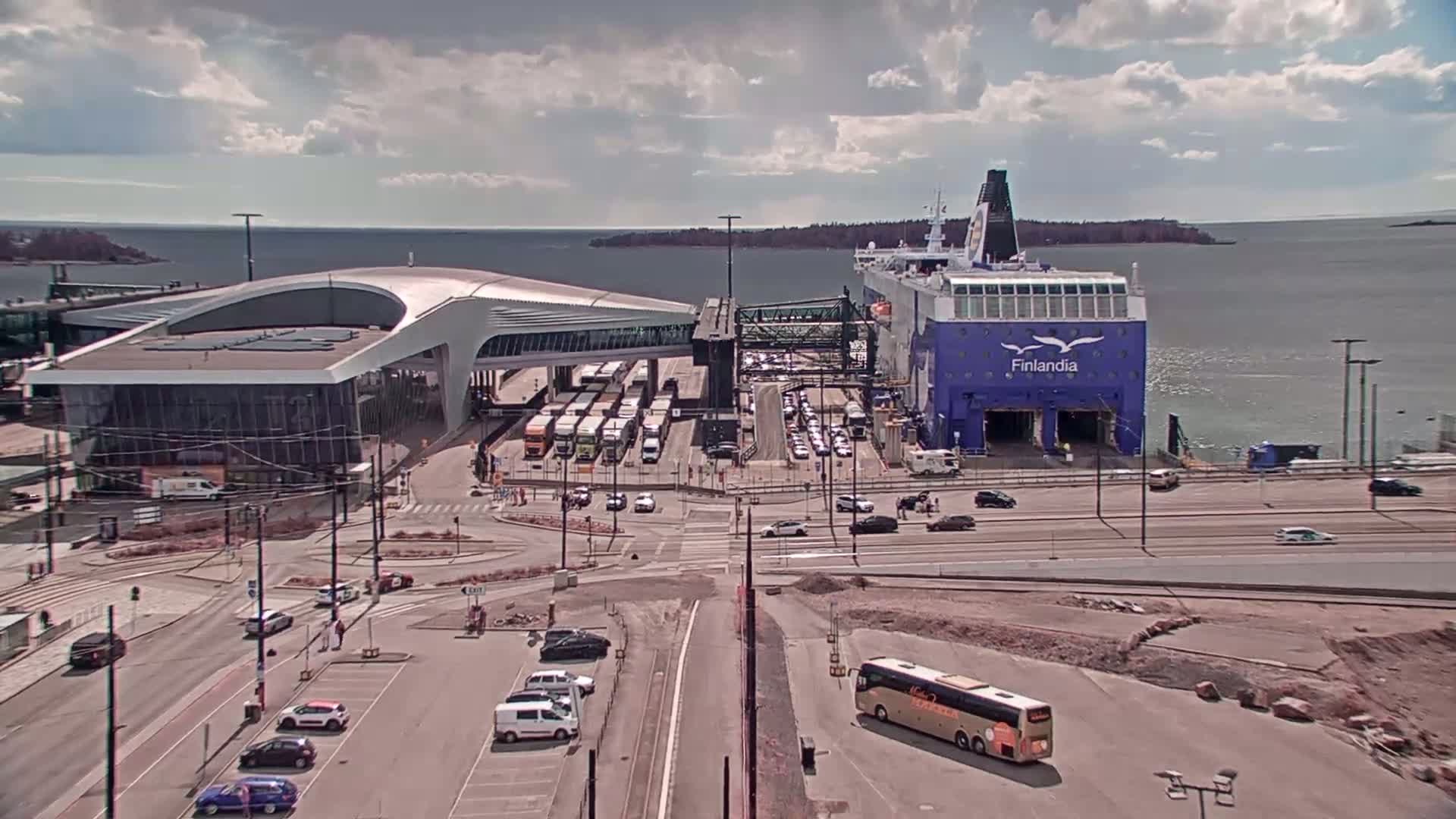 A large ferry is docked at a terminal building, with numerous vehicles parked nearby under partly cloudy skies.