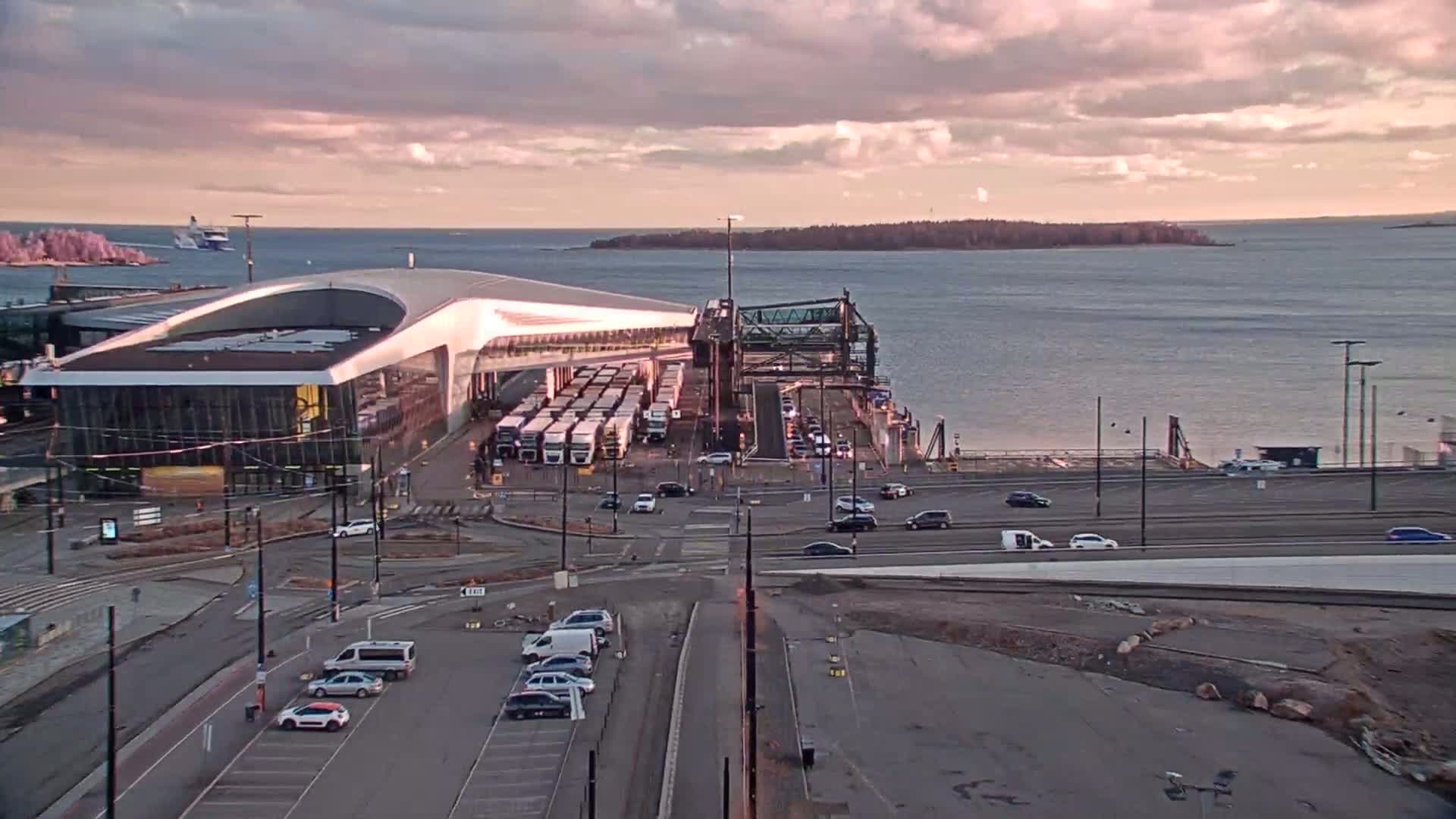 A ferry terminal with numerous vehicles, including buses and cars, is situated by a body of water under a cloudy, pinkish sky.