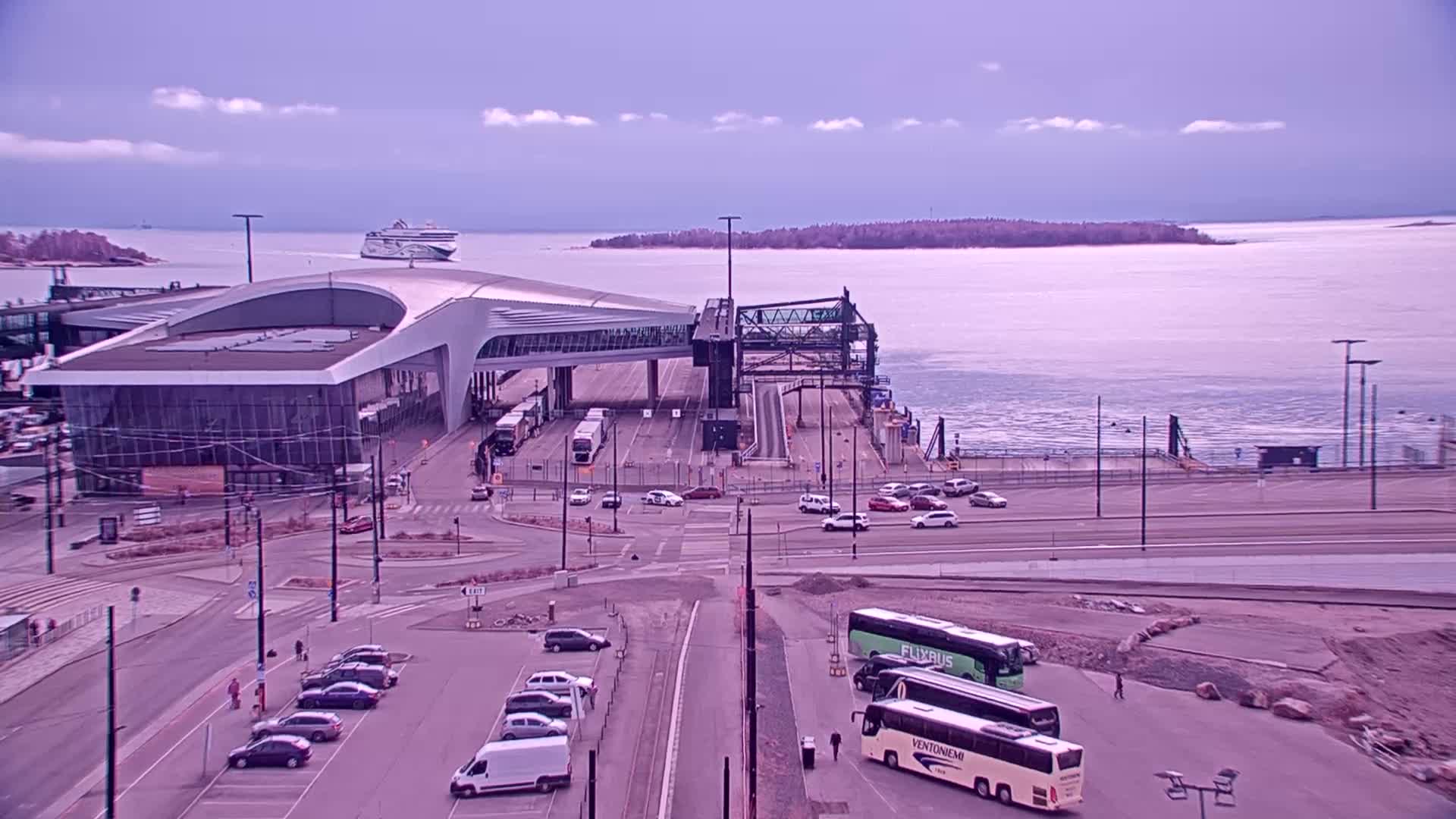 A ferry terminal with a modern building and several cars and buses in a parking lot, overlooks a calm body of water with a ferry in the distance under a cloudy sky.