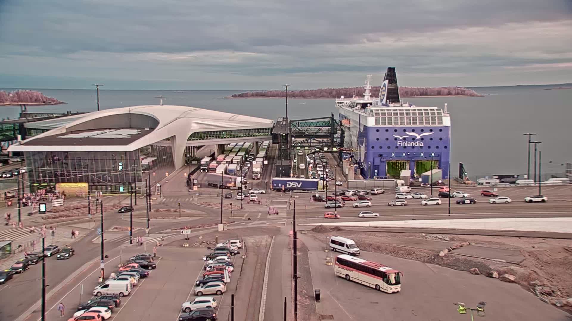 A large ferry is docked at a terminal, surrounded by cars, trucks, and buses under an overcast sky.