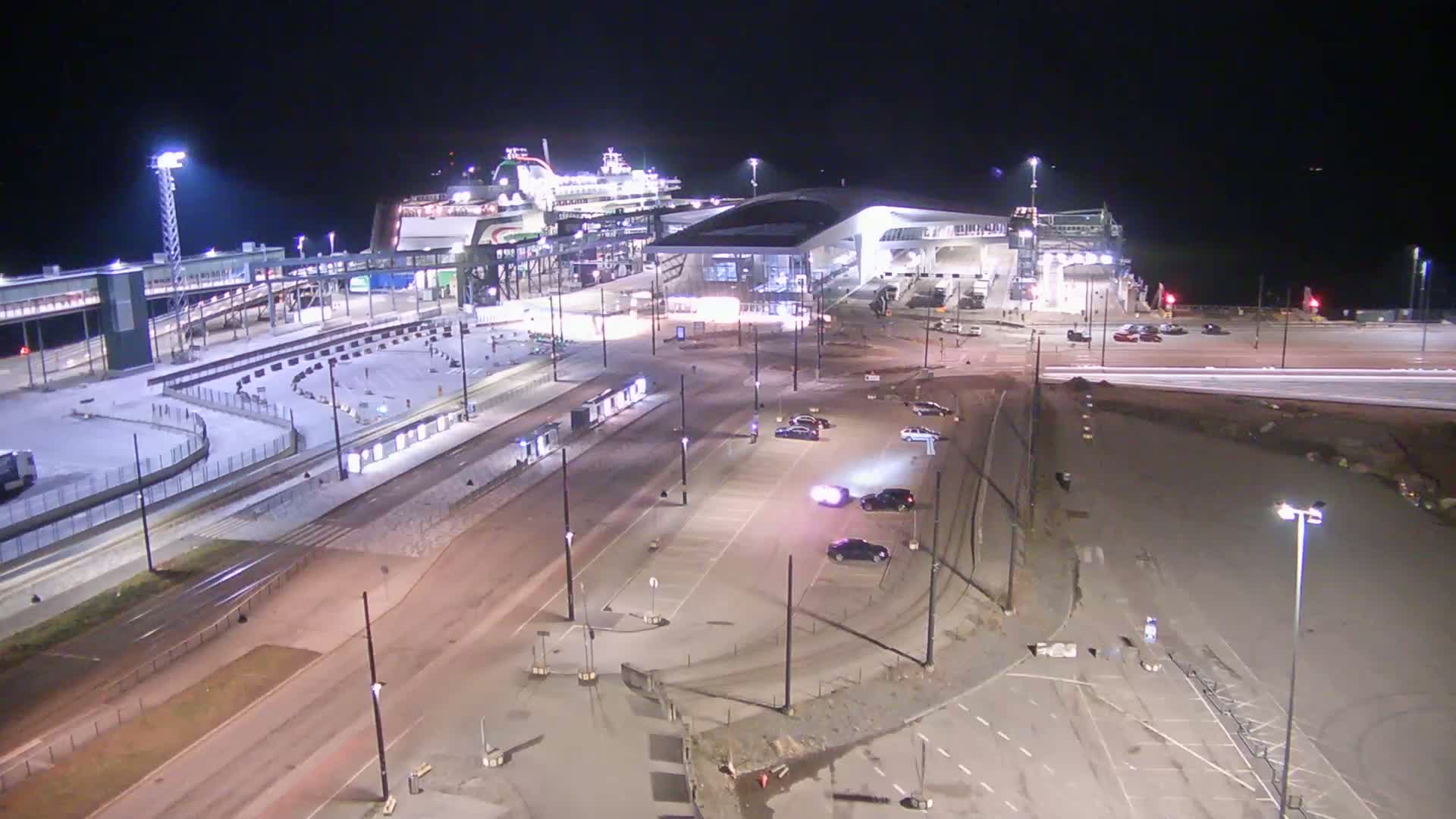 A ferry is docked at a terminal at night, with some cars parked in the surrounding lots, and light snow on the ground.