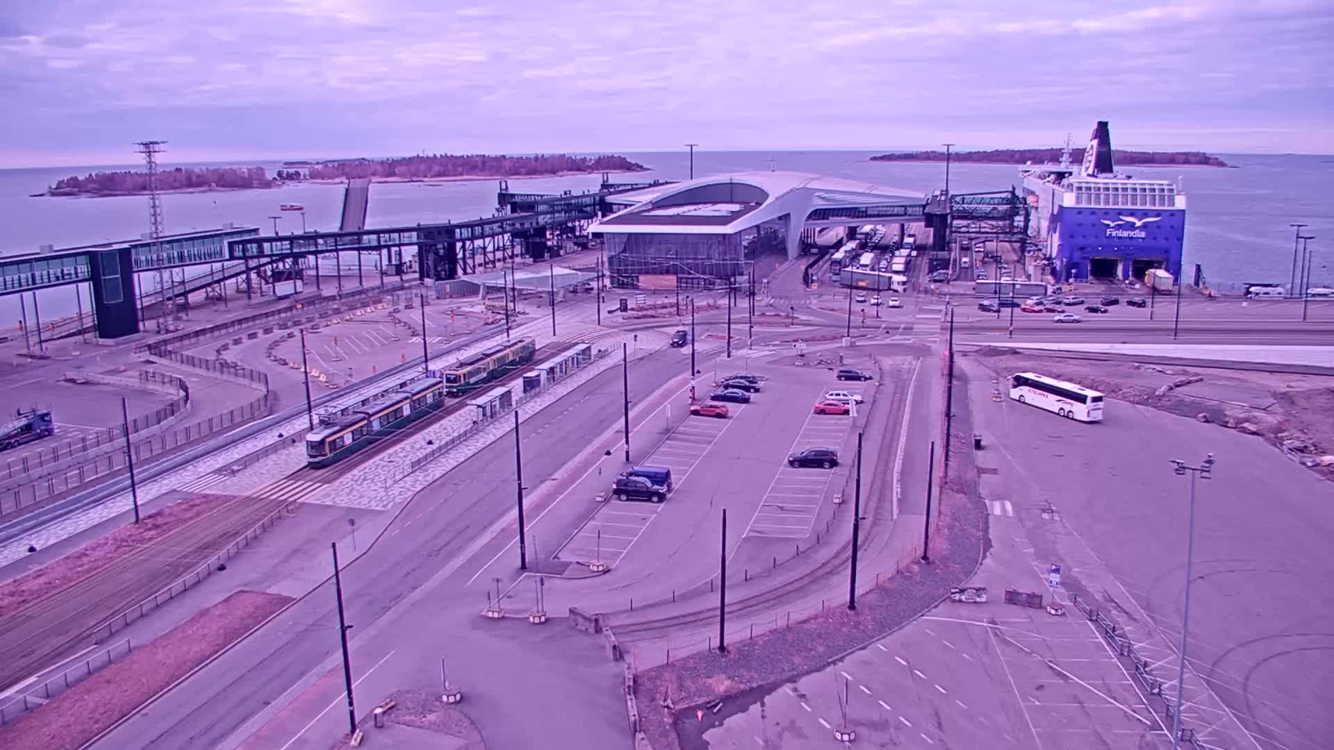 An aerial view shows a ferry terminal with a large ferry docked, trams on tracks, cars in a parking lot, and a bus, all under an overcast sky.