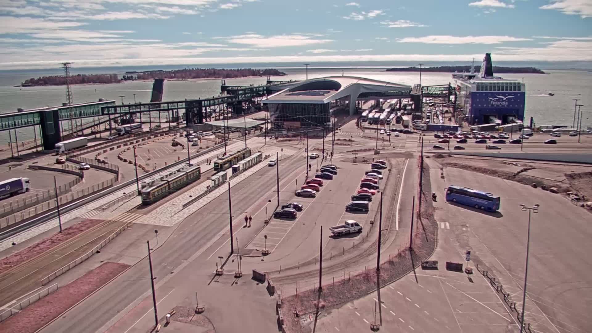 A ferry terminal on a sunny day with a large ferry docked, several cars parked, trams on the tracks, and a bus in a lot.