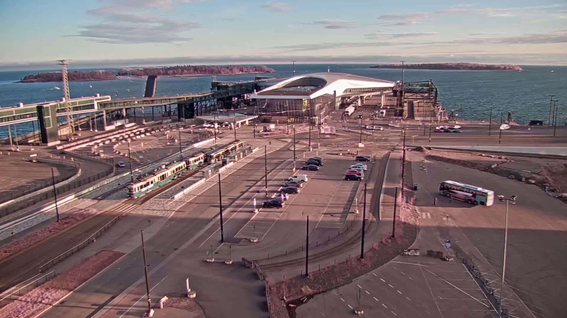 A ferry terminal with a train and several cars parked, under a mostly clear sky, next to calm water and small islands in the distance.