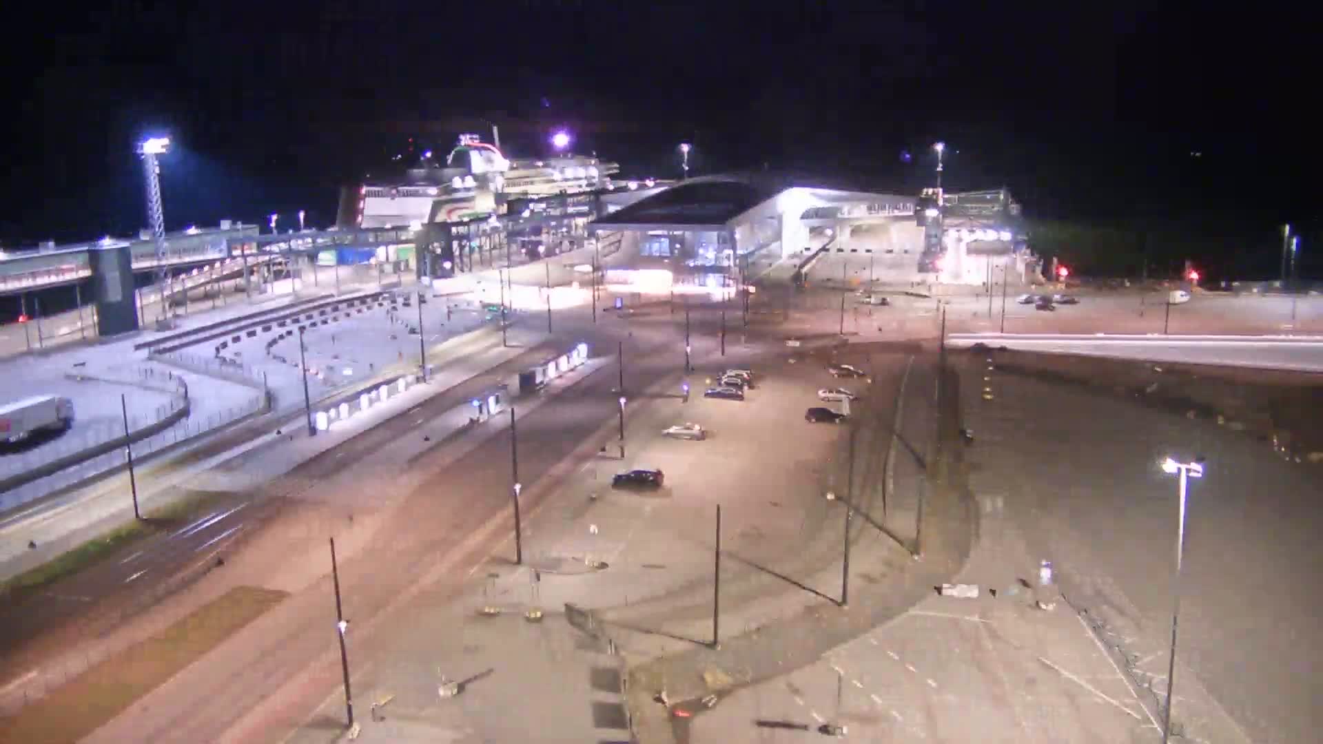 A large ferry is docked at a terminal at night, with several cars parked in nearby lots and roadways illuminated by streetlights.
