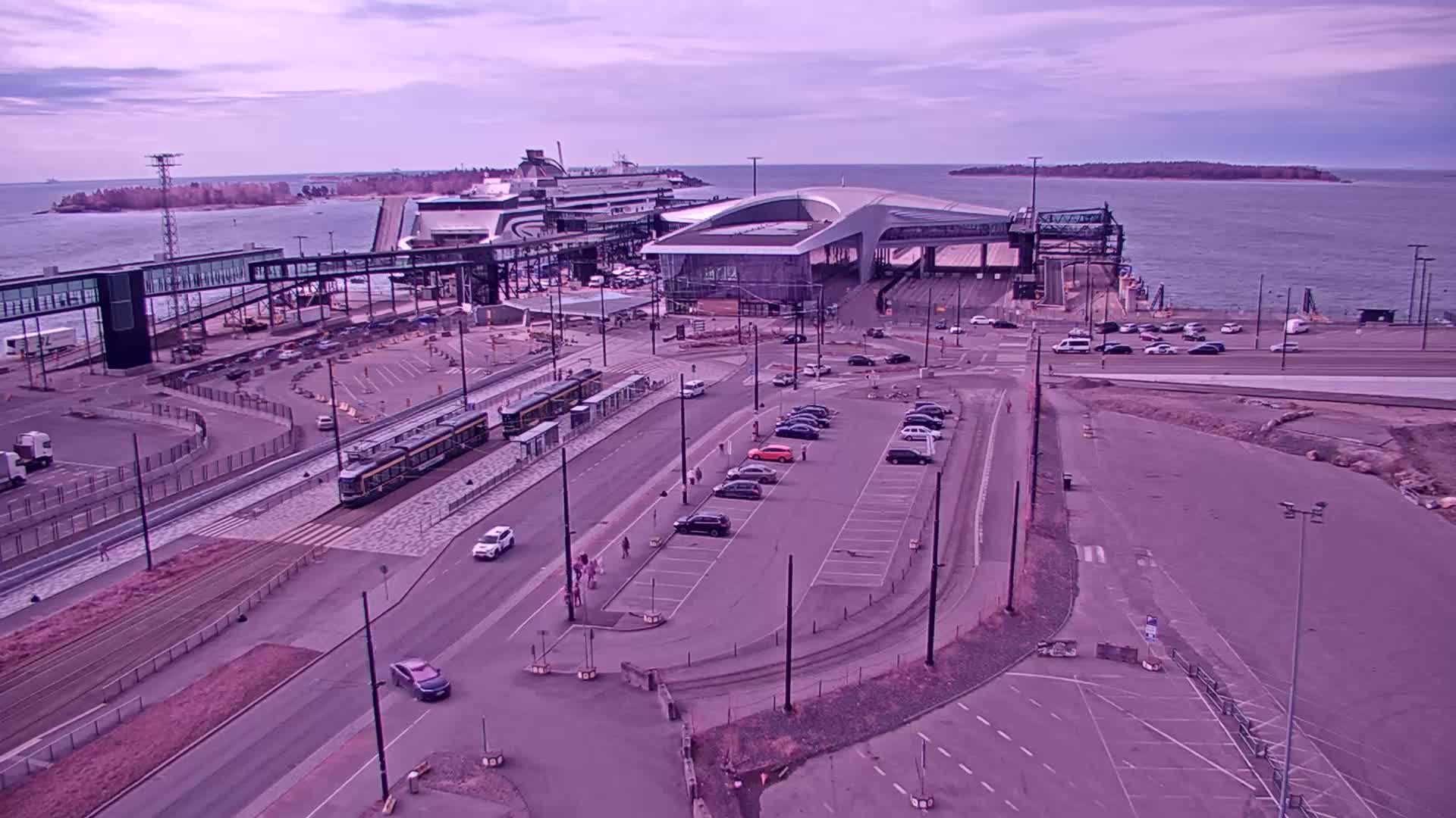 A ferry terminal with a tram and several cars parked in a lot, under a cloudy sky.