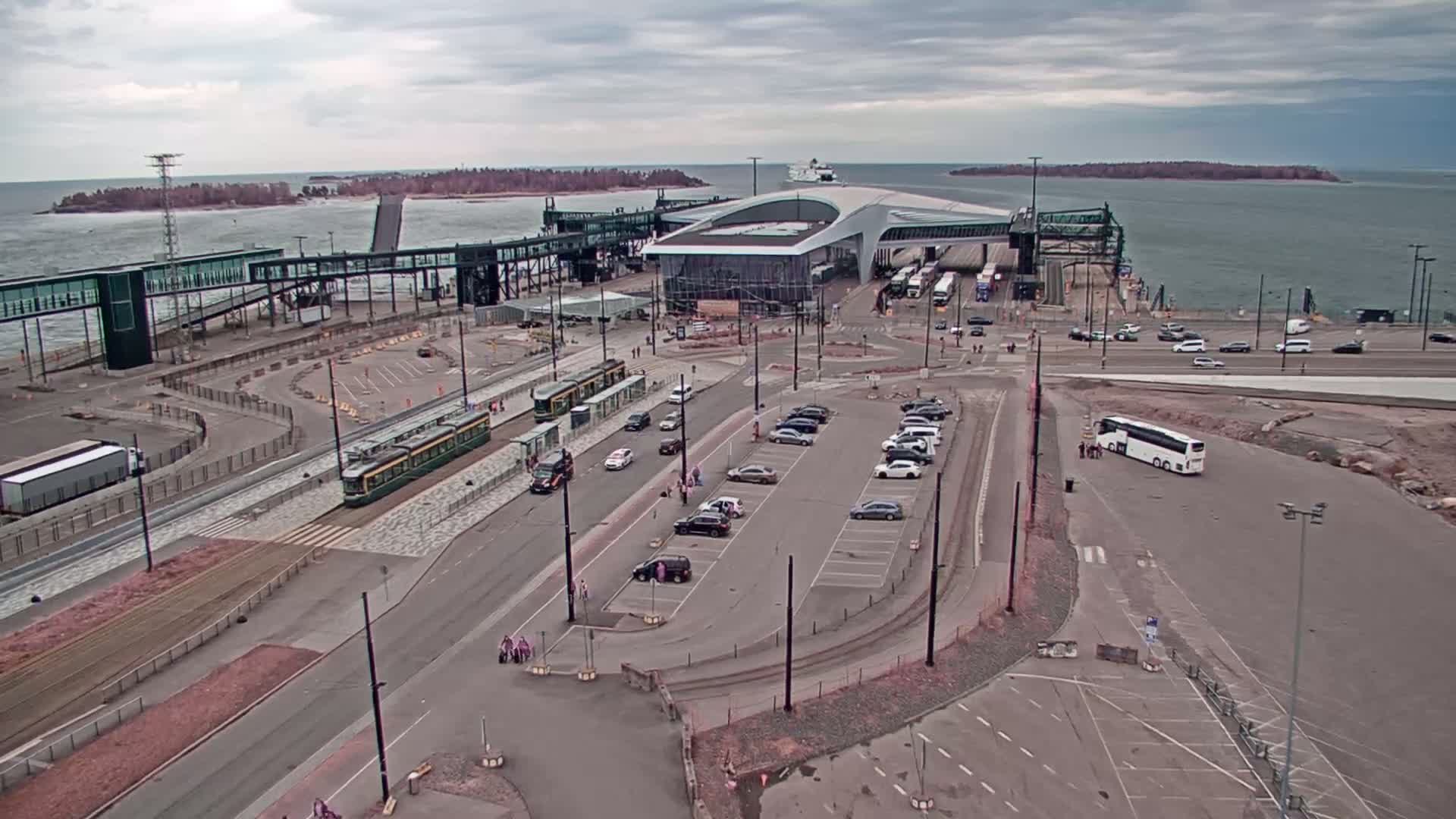 An aerial view shows a ferry terminal with a tram, cars, buses, and a ferry in the harbor on an overcast day.