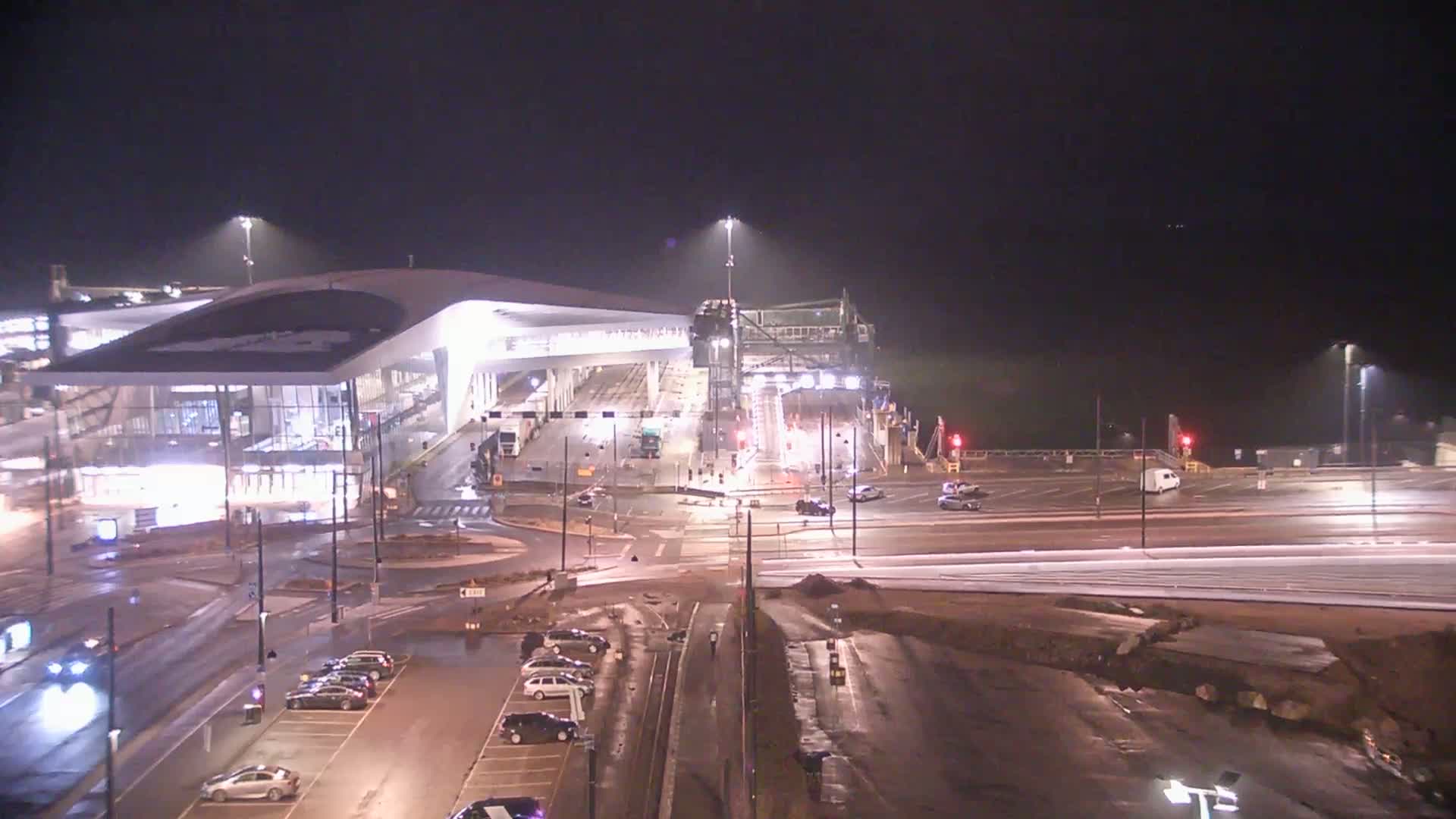 A nighttime, wet scene shows a modern ferry terminal with vehicles parked nearby and a ferry partially visible under a loading gantry.