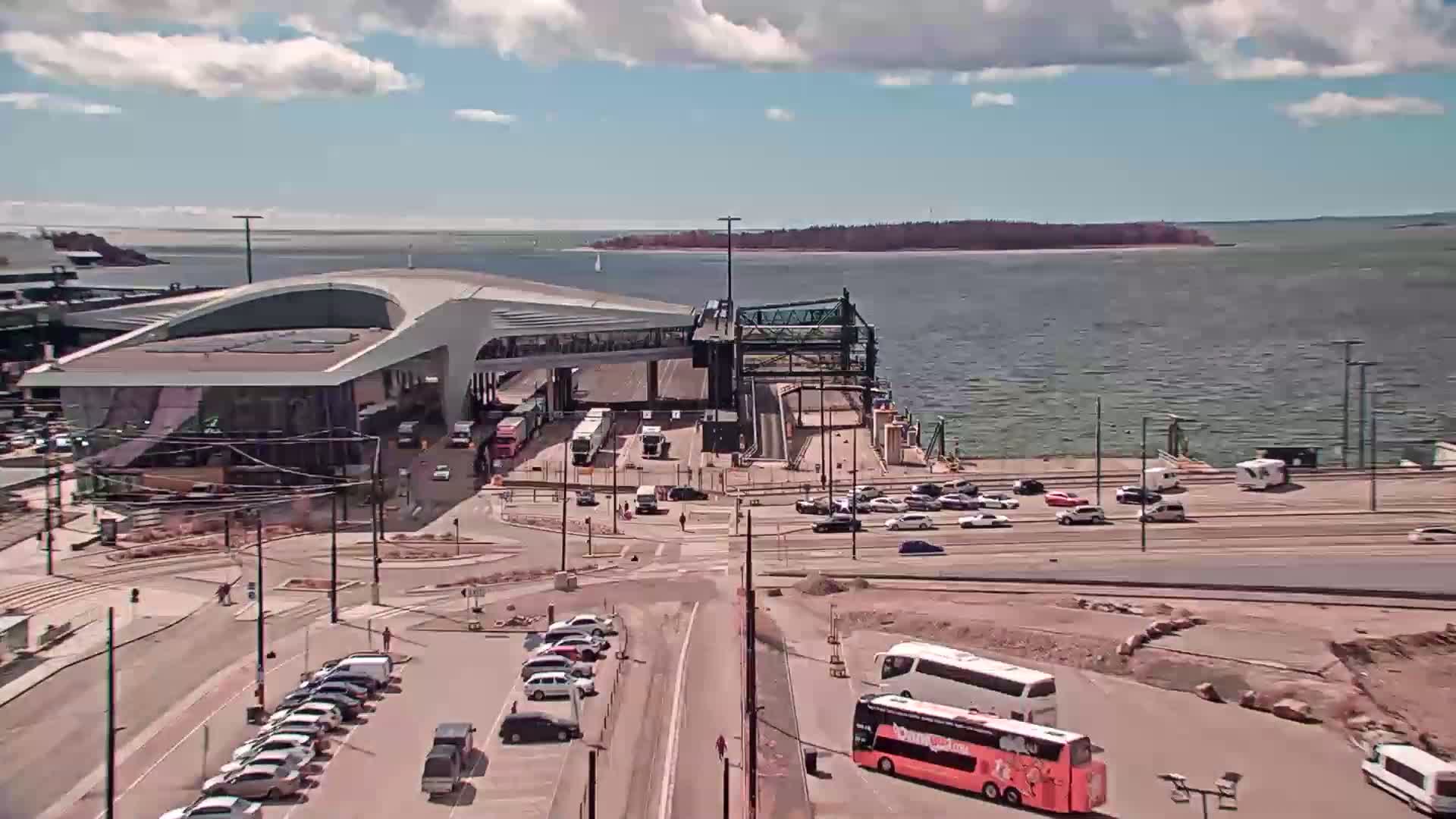 A ferry terminal with numerous vehicles parked and driving around it, on a partly sunny day next to a body of water.