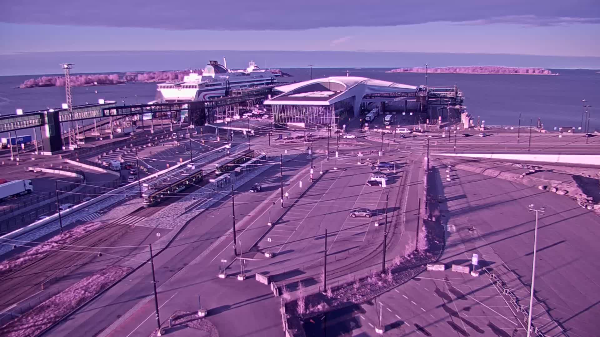 A ferry terminal with a large cruise ship docked, a tram on the road, and several cars parked in a lot, all under a clear, bright sky.