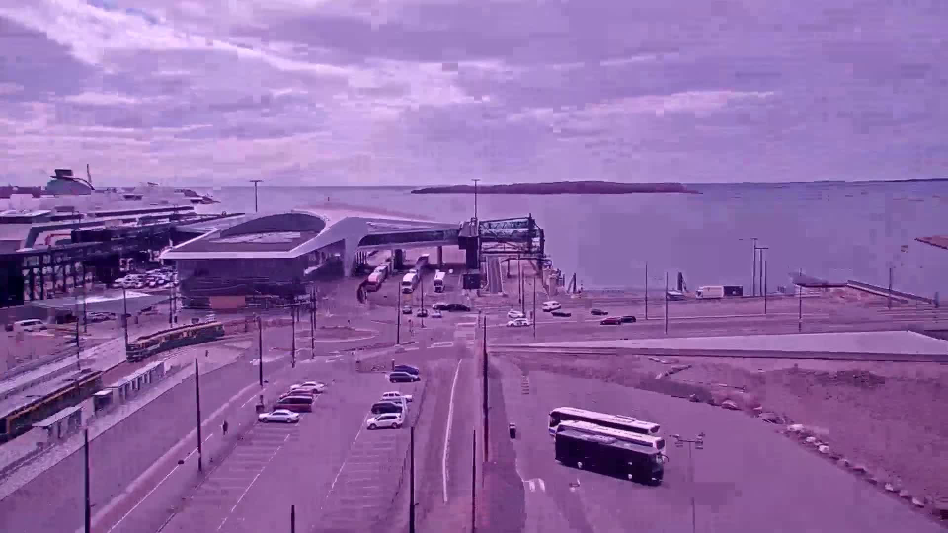 A ferry terminal with several vehicles and a bus, near a body of water under a partly cloudy sky.