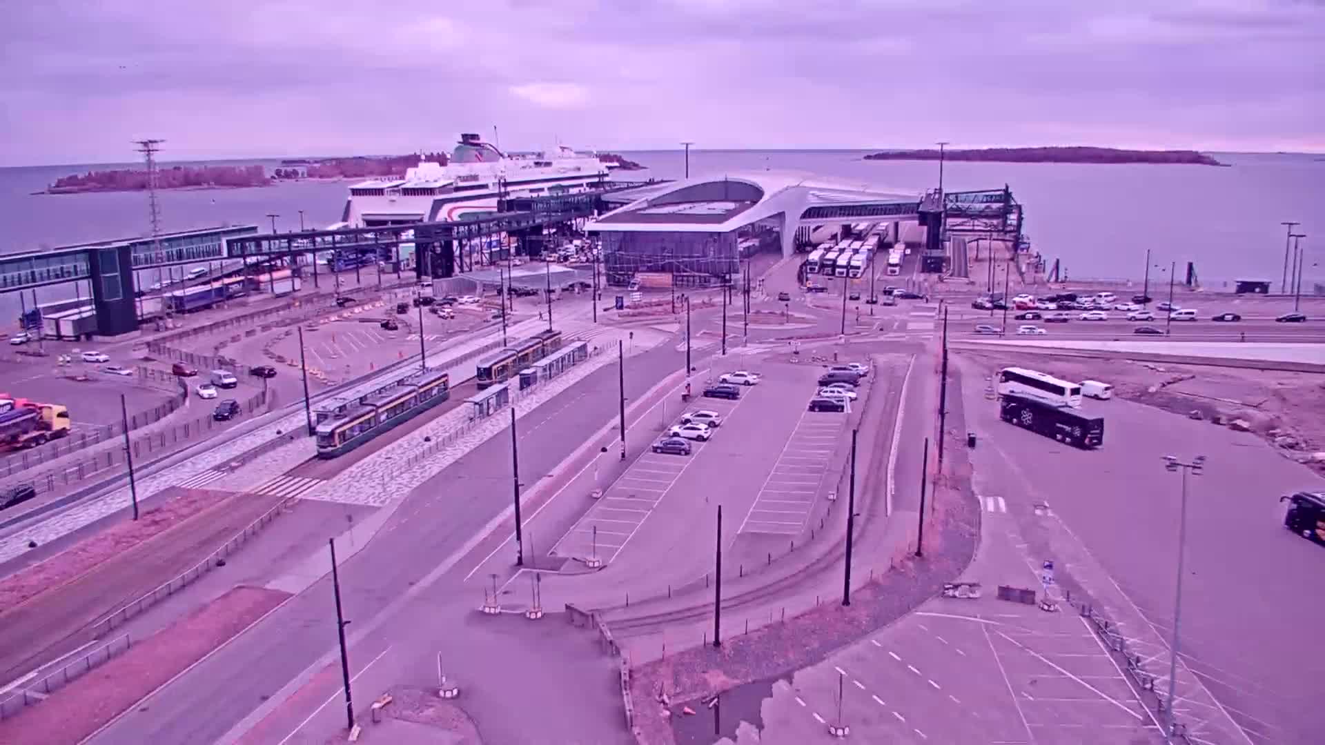 An aerial view shows a ferry terminal with trams, cars, buses, and a ferry docked under an overcast sky.