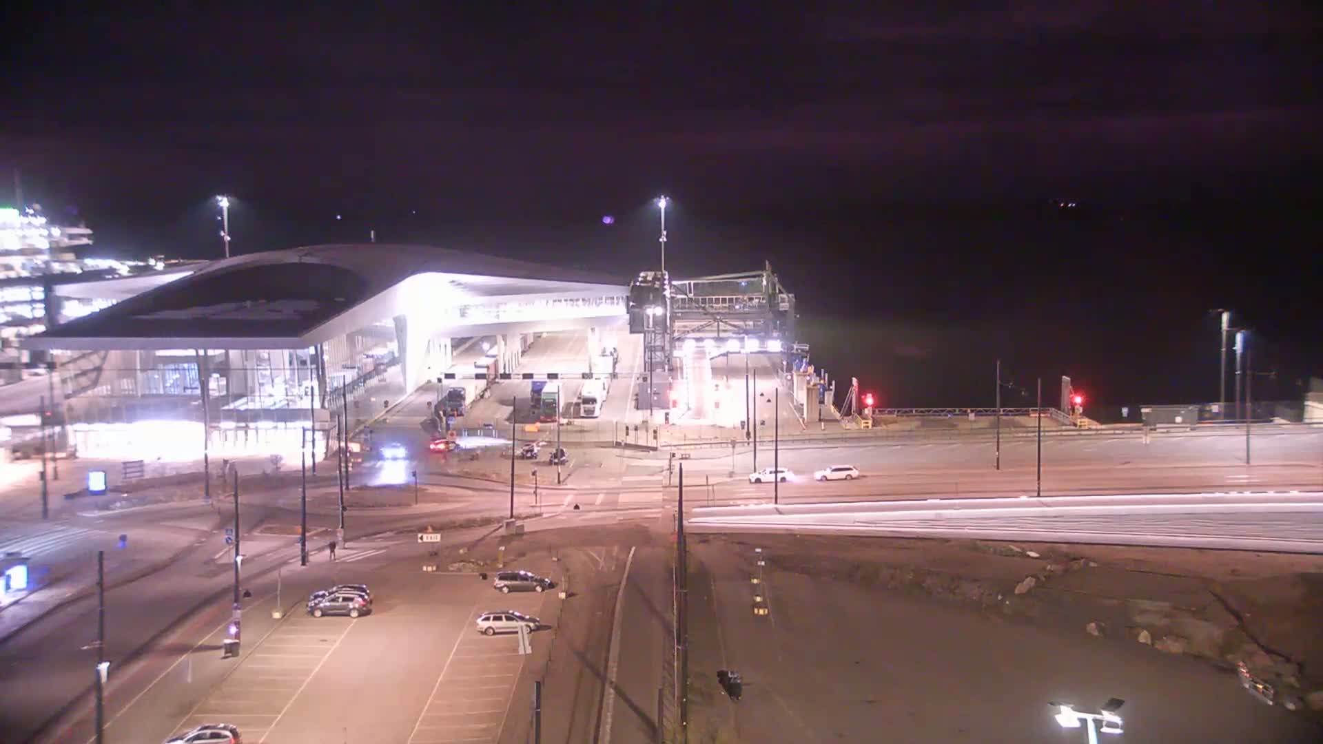 A brightly lit ferry terminal at night, with several cars in a nearby parking lot and some traffic on roads leading to the terminal.