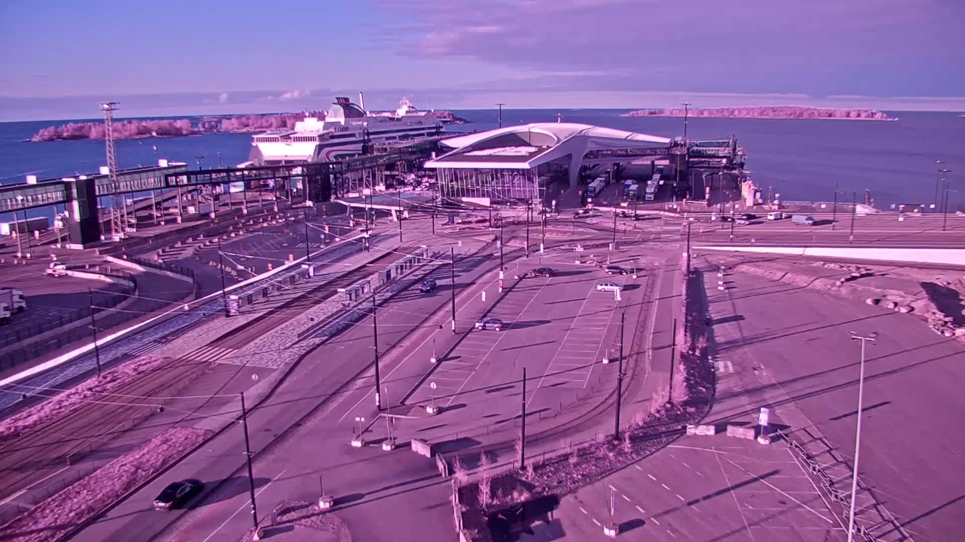 A ferry terminal with a large ferry docked, a modern terminal building, and a mostly empty parking lot, all under a clear, sunny sky.