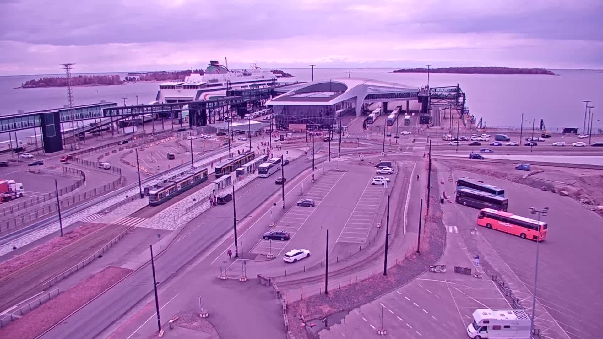 An aerial view shows a ferry terminal with a docked ferry, trams, buses, cars, and pedestrians under an overcast sky.