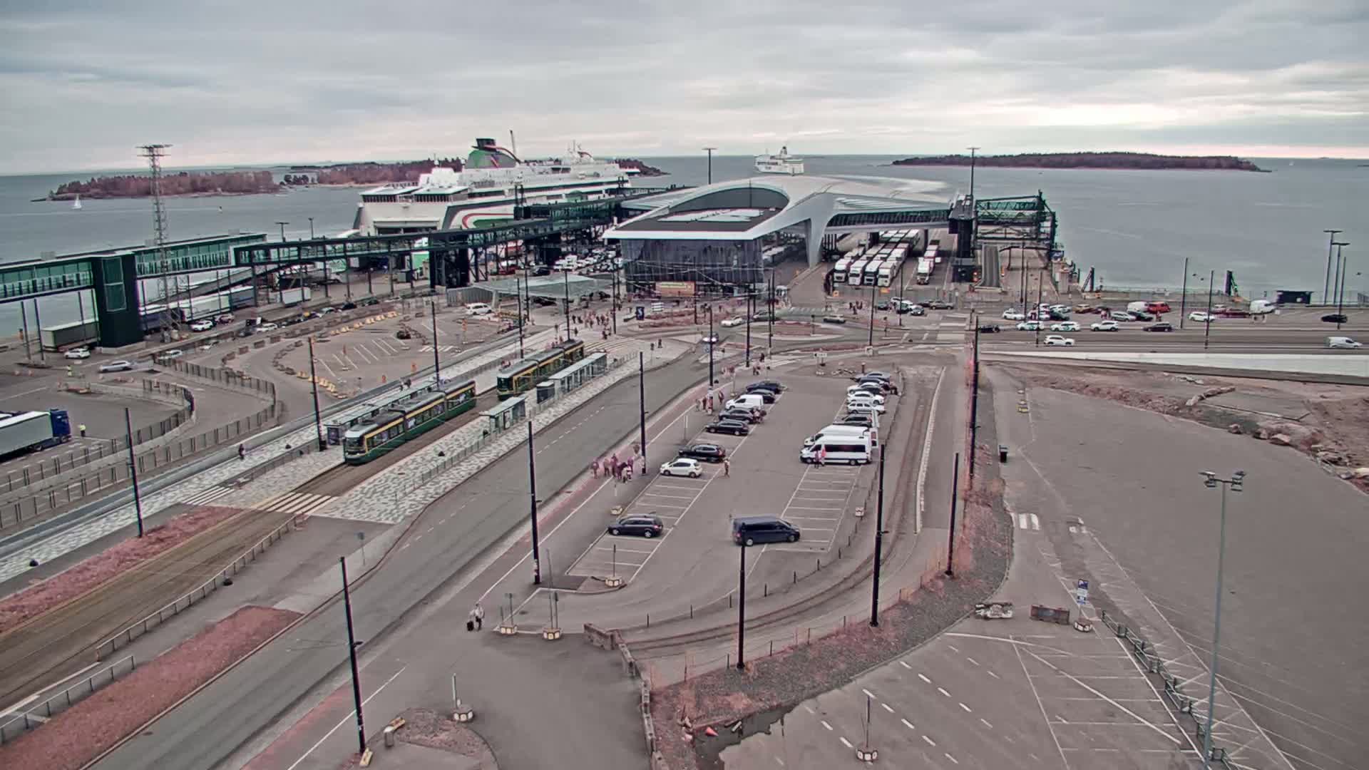An aerial view of a ferry terminal on an overcast day shows two ferries docked, trams at a station, cars in a parking lot, and buses near the terminal building.