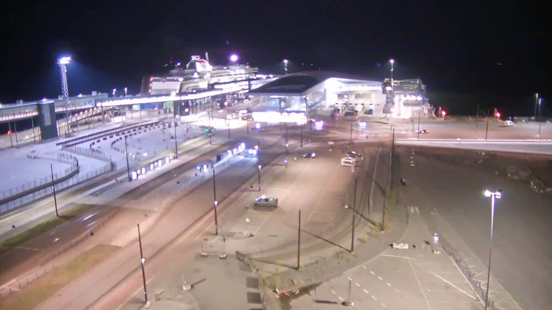 A brightly lit ferry terminal at night, with a large ferry docked and several vehicles in a nearby parking lot.
