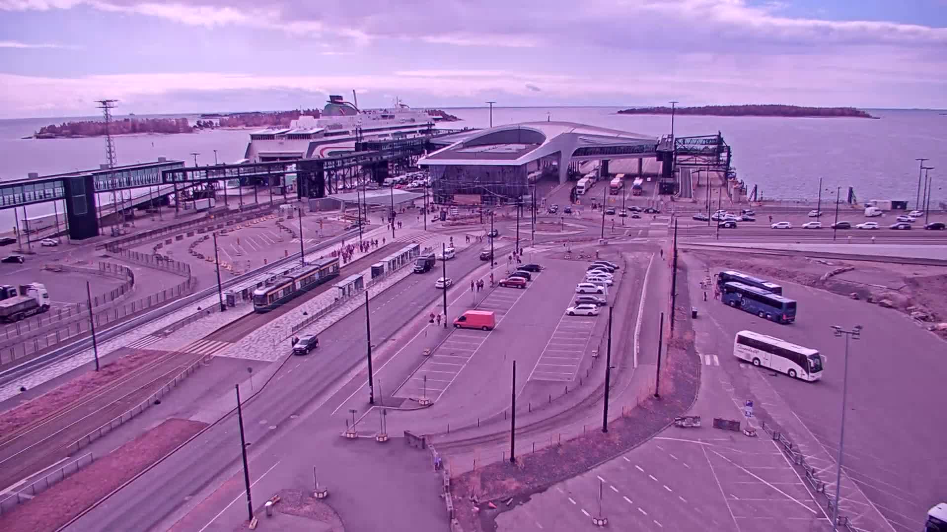 An aerial view of a ferry terminal with a docked ferry, buses, cars, and a tram, under a cloudy sky.