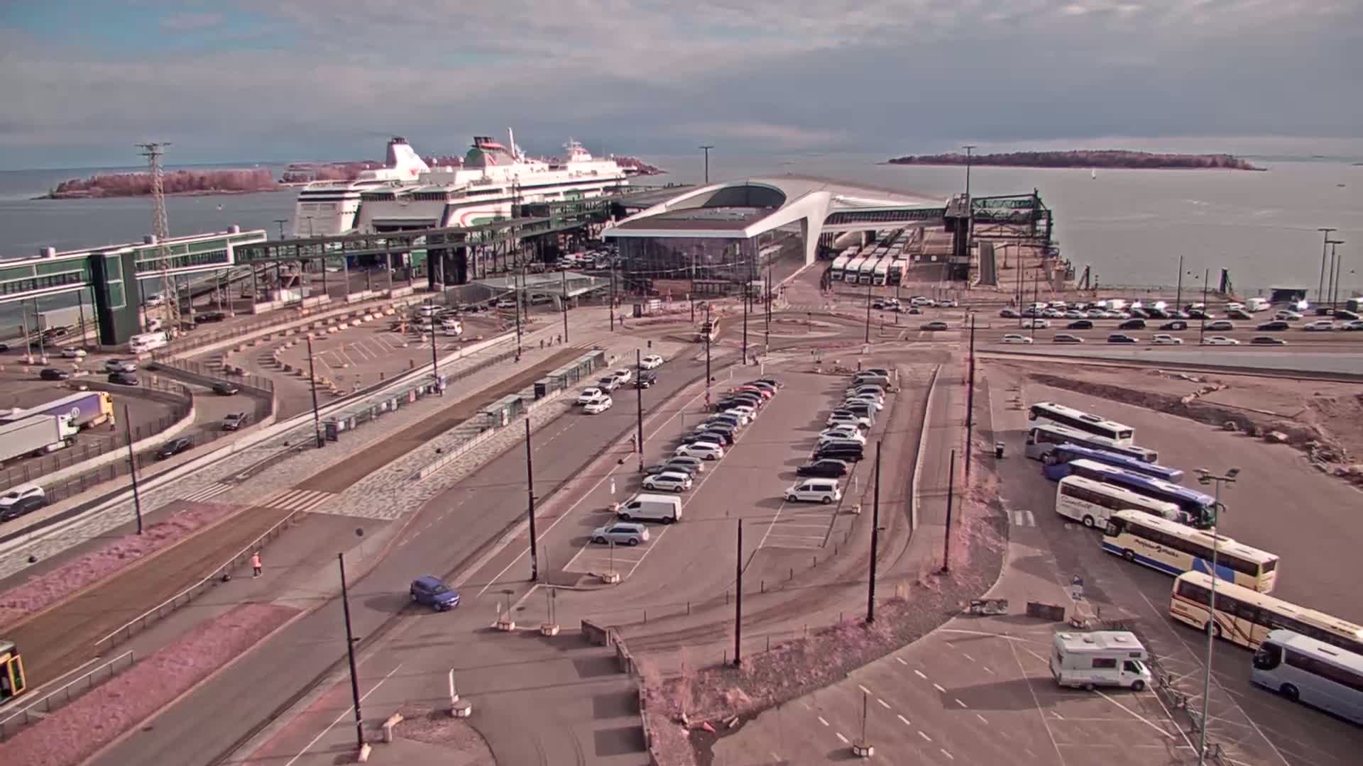 An aerial view shows a ferry docked at a terminal, with cars, buses, and a few pedestrians in the surrounding parking lots and roadways on a partly cloudy day.