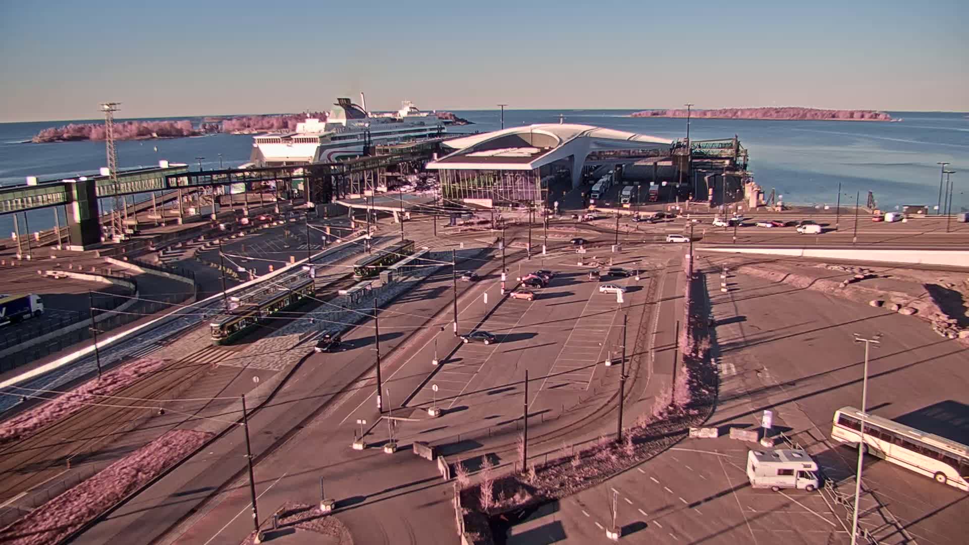 A ferry terminal with trams and vehicles in a parking lot on a sunny day, overlooking a calm body of water with islands in the distance.