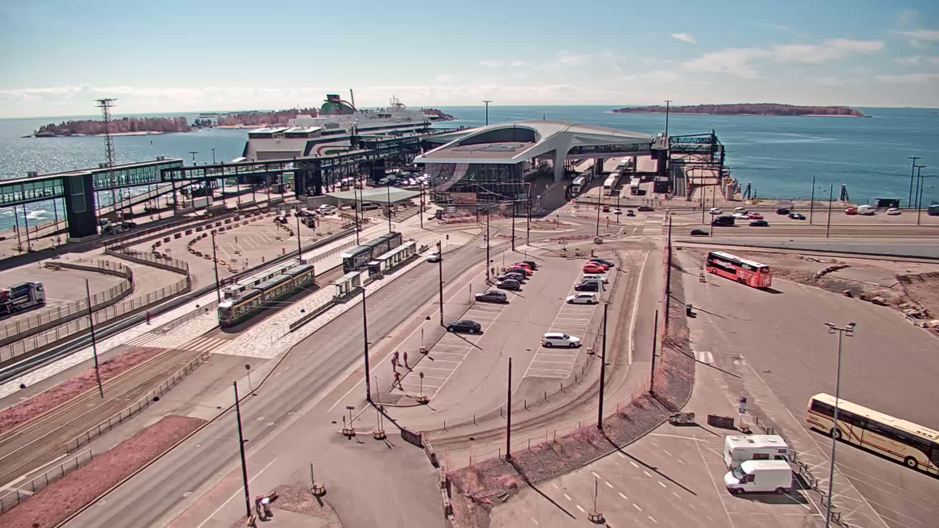 A ferry terminal on a sunny day with a tram, buses, and cars in the parking lot, alongside a body of water with islands in the distance.