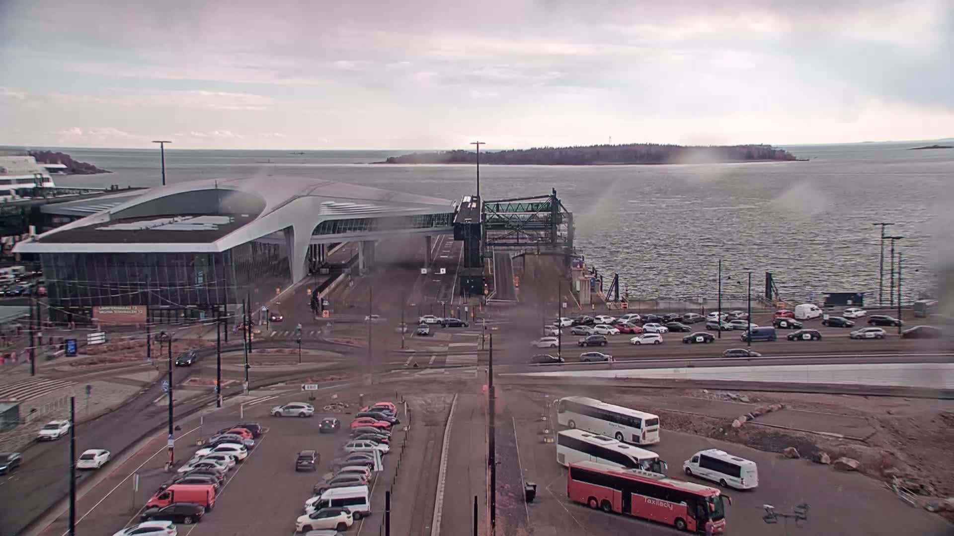A ferry terminal with a modern building and numerous cars and buses parked nearby, on an overcast day.