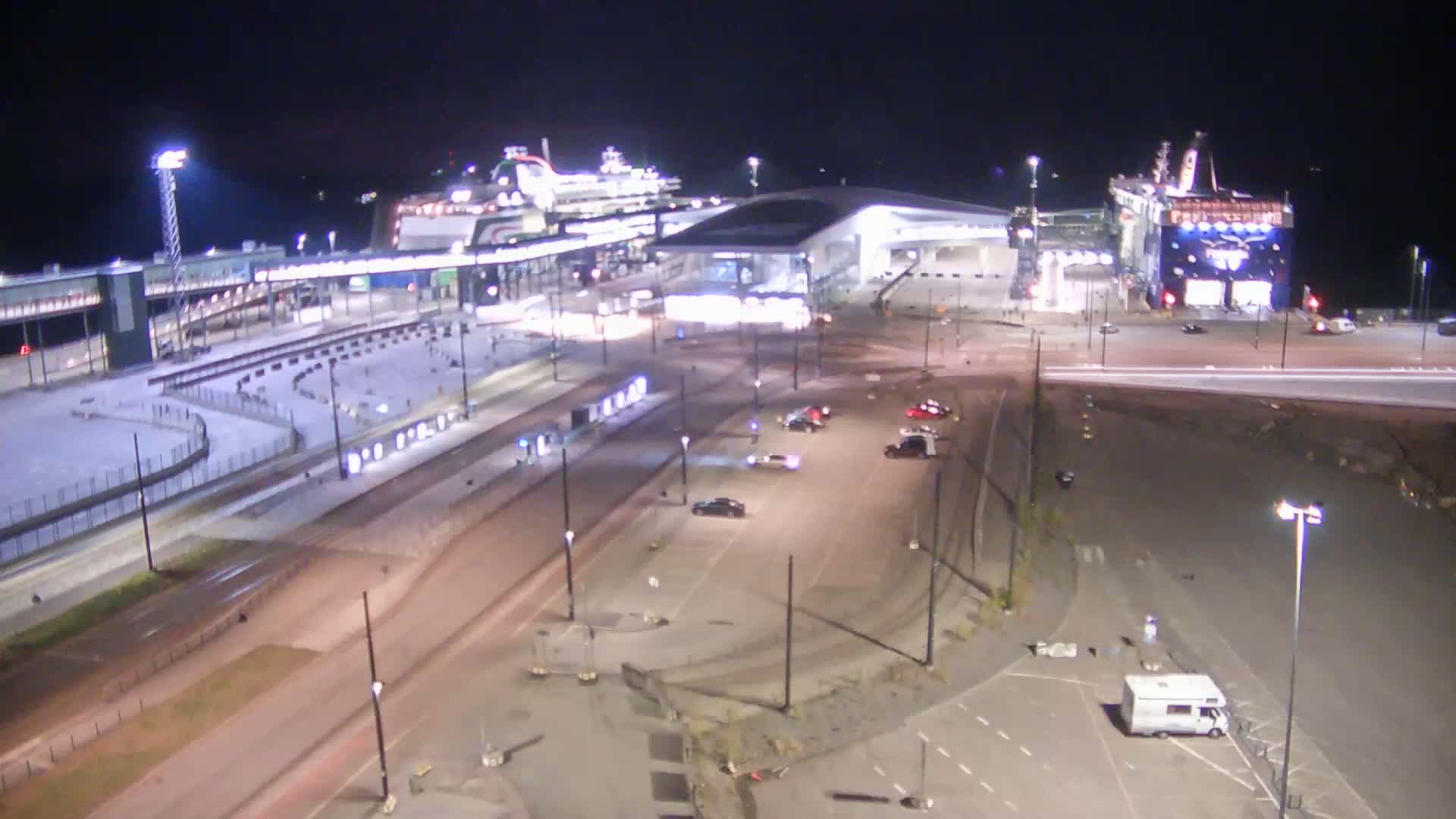 At night, a ferry terminal is brightly lit, with two large ferries docked and several cars parked in the surrounding lots.