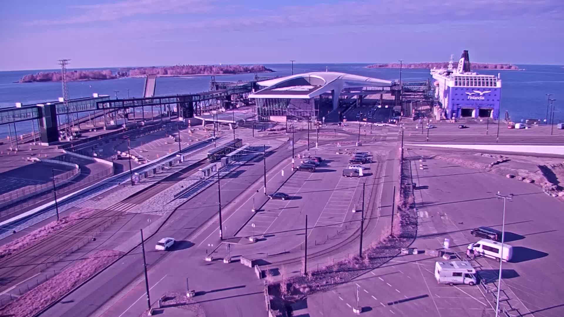 An aerial view shows a ferry terminal with a large ferry docked,  tram tracks, parking lots, and a walkway over the water, on a sunny day.